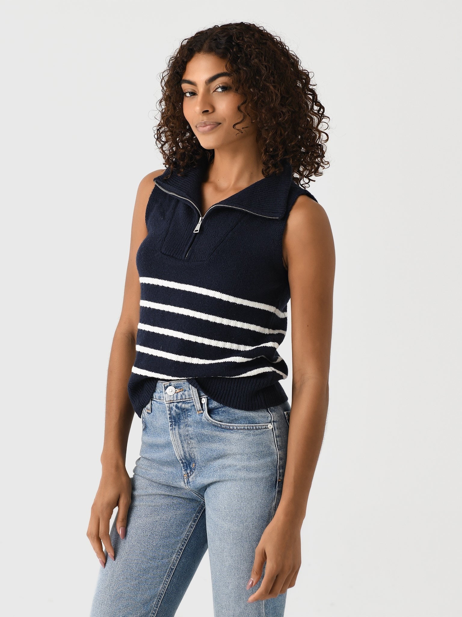 A woman with curly hair is wearing the Matthew Bruch Quarter Zip Navy Knit Striped Knit Nest sweater, made of recycled nylon and featuring white horizontal stripes across the middle. She pairs it with light blue jeans and stands against a plain background, posing with her left hand resting on her thigh.