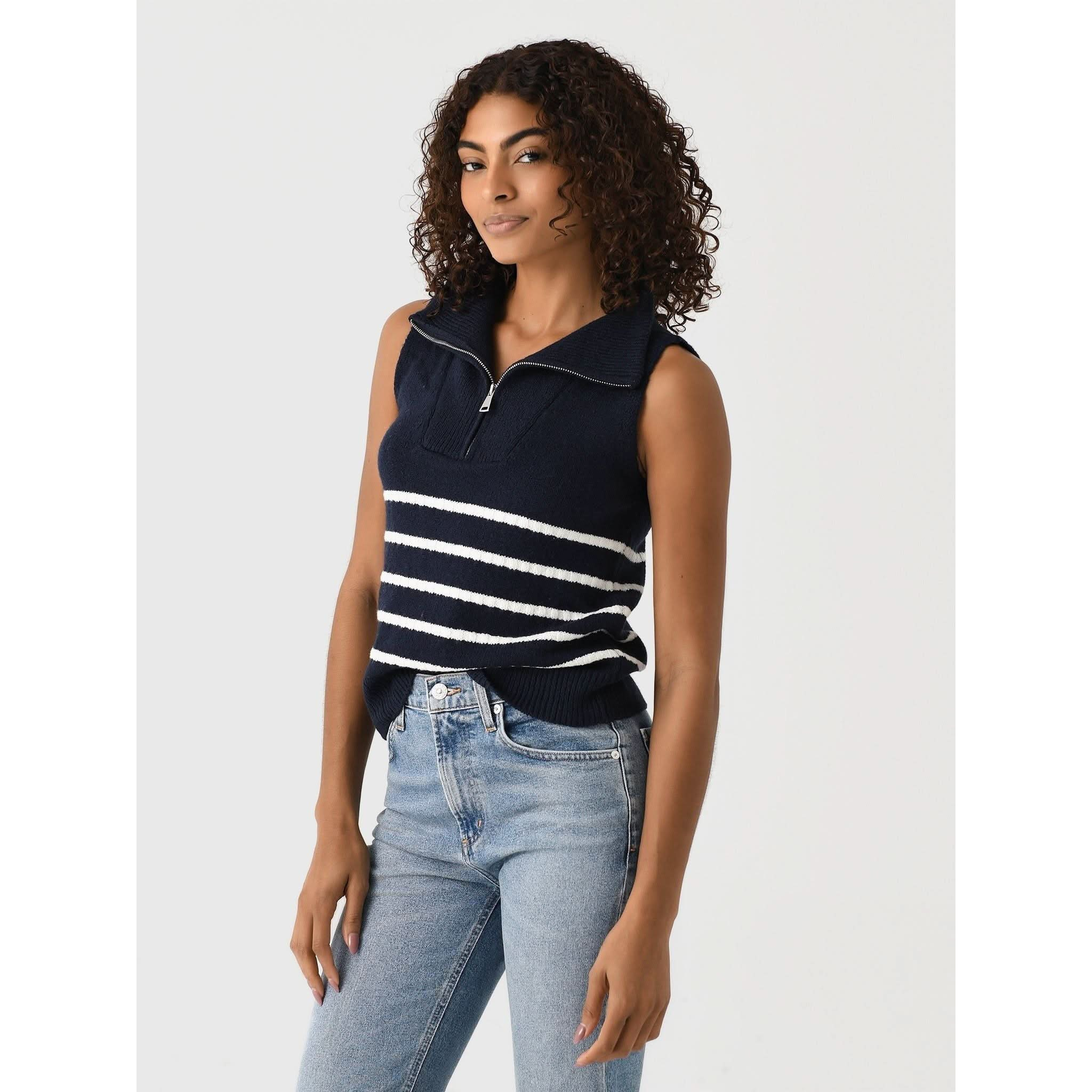 A woman with curly hair is wearing the Matthew Bruch Quarter Zip Navy Knit Striped Knit Nest sweater, made of recycled nylon and featuring white horizontal stripes across the middle. She pairs it with light blue jeans and stands against a plain background, posing with her left hand resting on her thigh.