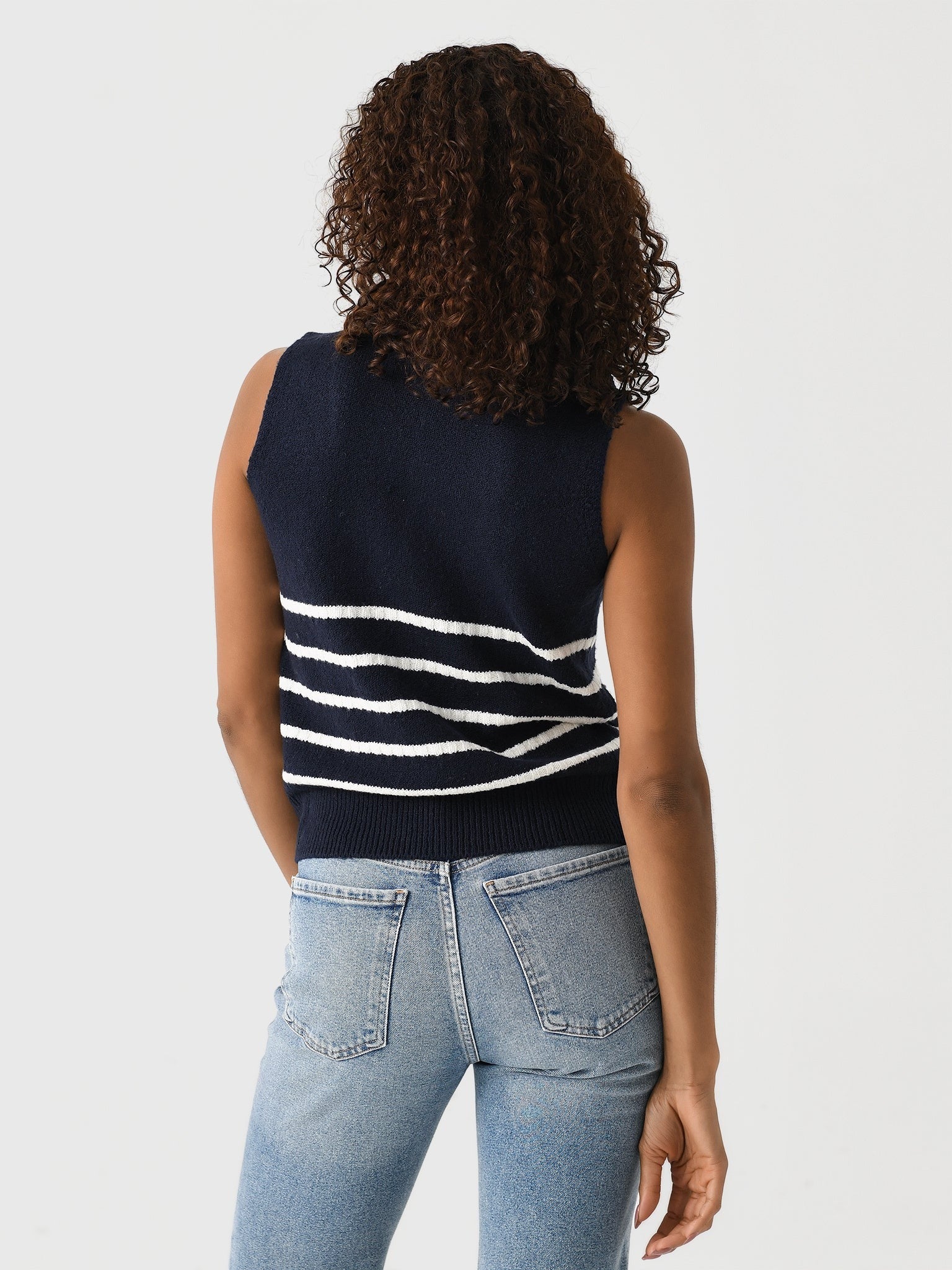 A person with curly hair stands facing away from the camera, wearing the Matthew Bruch Quarter Zip Navy Knit Striped Knit Nest, a sleeveless sweater made of merino wool with white horizontal stripes on the back. They are also sporting light blue jeans against a plain, white wall background.