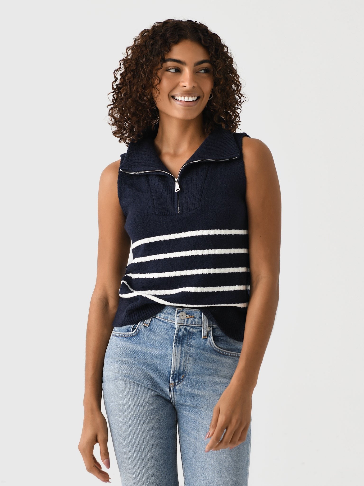 A person with curly hair is smiling and wearing the Matthew Bruch Quarter Zip Navy Knit Striped Knit Nest top, made of recycled nylon, paired with faded blue jeans. The background is plain and light-colored.