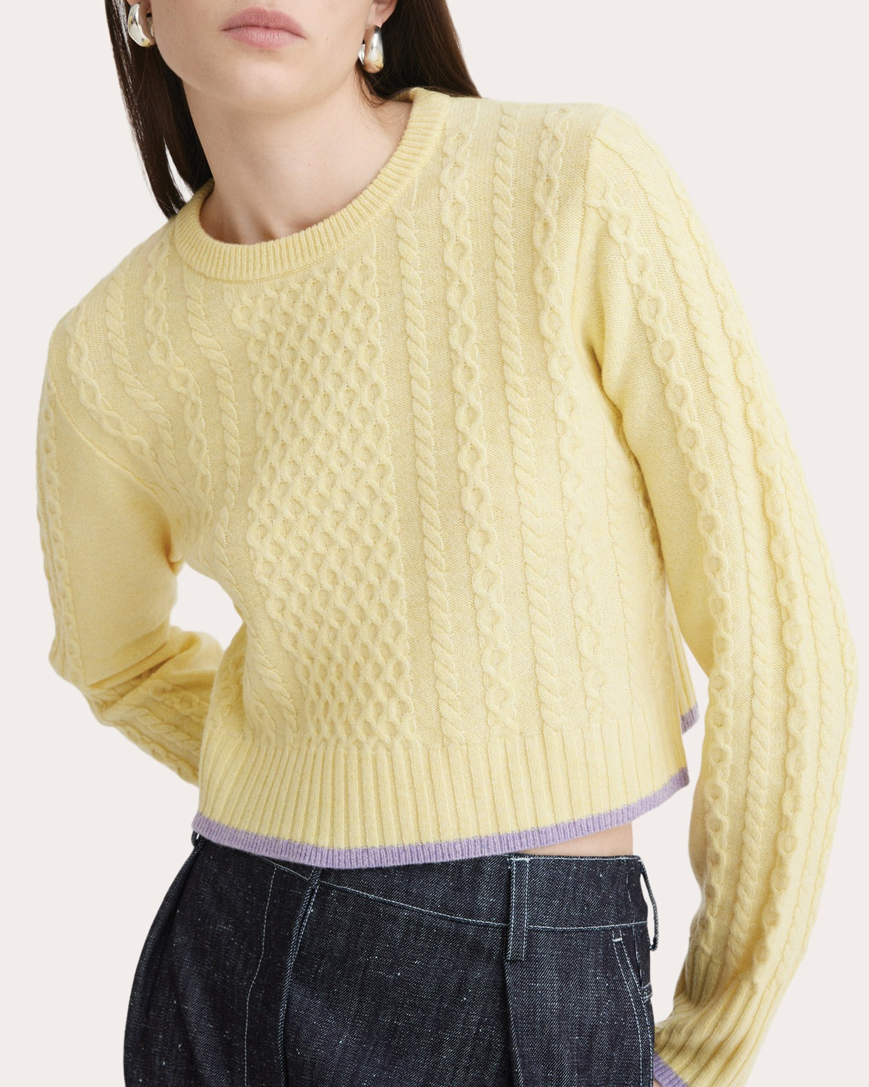 A person wearing a Matthew Bruch Cable Knit Crew Neck Sweater in light yellow, crafted from merino wool with ribbed cuffs and hem, paired with dark denim jeans. The hem of the sweater features a light purple accent. The person is posing with one hand on their hip.