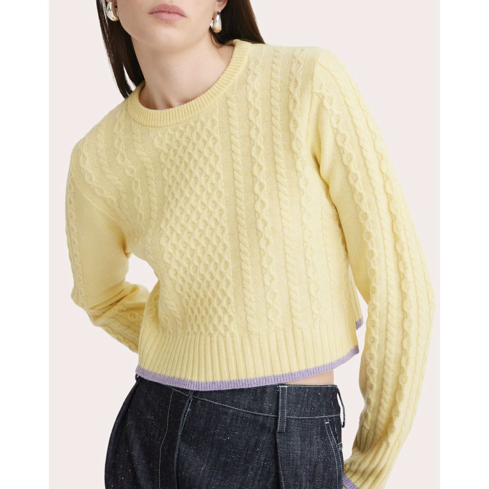 A person wearing a Matthew Bruch Cable Knit Crew Neck Sweater in light yellow, crafted from merino wool with ribbed cuffs and hem, paired with dark denim jeans. The hem of the sweater features a light purple accent. The person is posing with one hand on their hip.