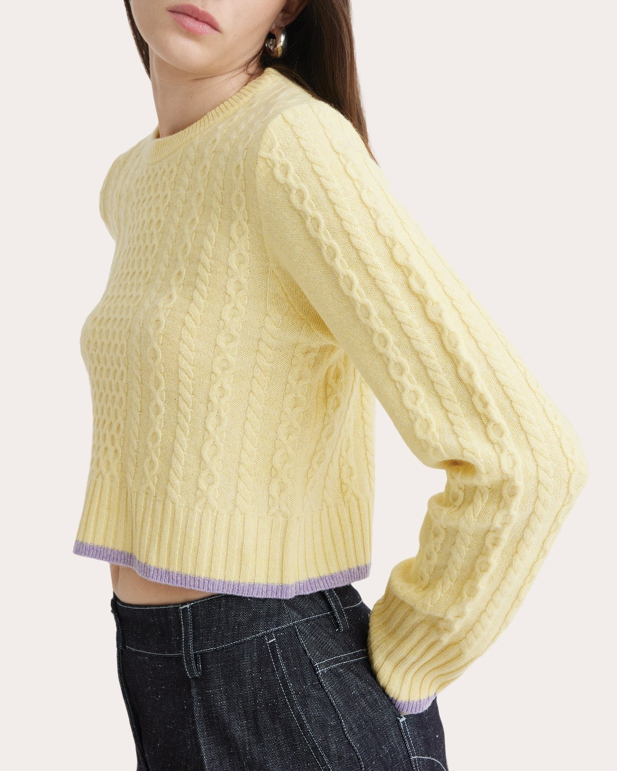 A person wearing the Matthew Bruch Cable Knit Crew Neck Sweater in pastel yellow, made from recycled nylon with intricate cable knit designs, paired with high-waisted navy blue jeans. The sweater features a subtle light purple trim at the hem and cuffs. The person is shown from the waist up, slightly turned to the side.