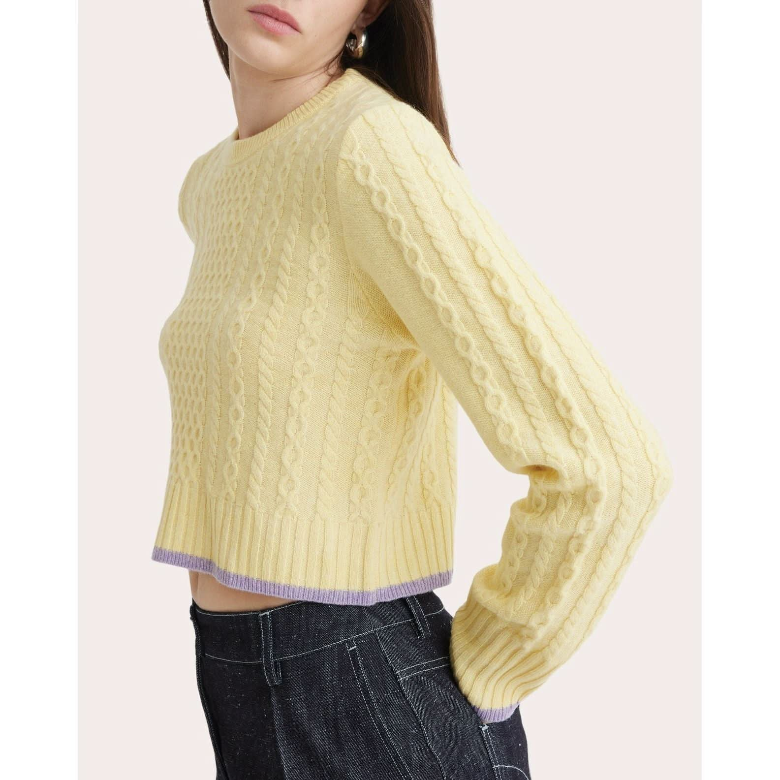 A person wearing the Matthew Bruch Cable Knit Crew Neck Sweater in pastel yellow, made from recycled nylon with intricate cable knit designs, paired with high-waisted navy blue jeans. The sweater features a subtle light purple trim at the hem and cuffs. The person is shown from the waist up, slightly turned to the side.