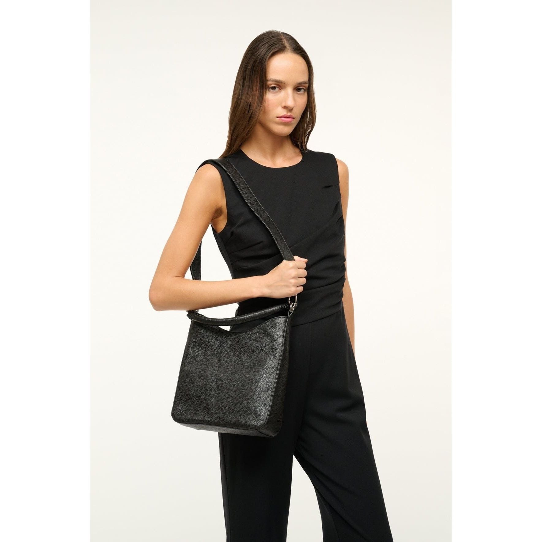 A person wearing a sleeveless black outfit is standing against a plain background, showcasing the sleek Staud Mel Bag in black cow leather, featuring a removable crossbody strap.