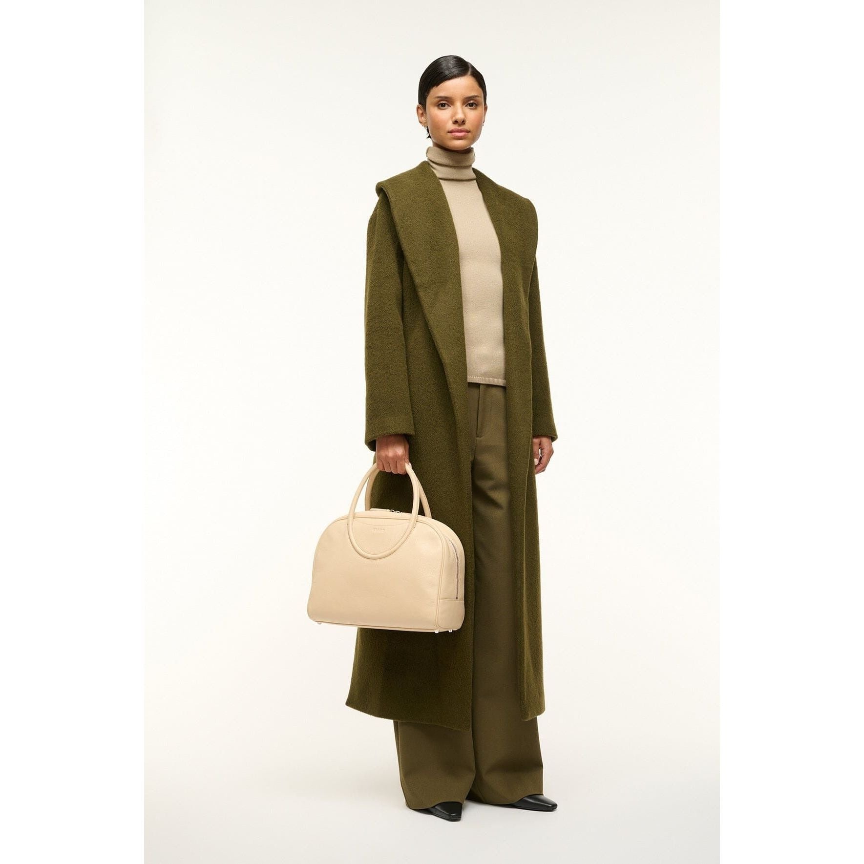 A person stands against a white background wearing a long olive green coat, beige turtleneck, and brown pants, holding a Staud Maude Bowler Bag in black polished leather with adjustable handles adorned with a Moon buckle.