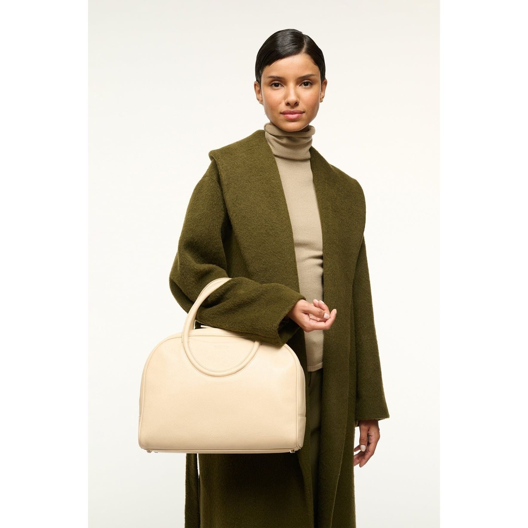 A person wearing a long, dark green coat and a beige turtleneck sweater holds the Staud Maude Bowler Bag in black, featuring polished leather with adjustable handles. The background is plain and light-colored.