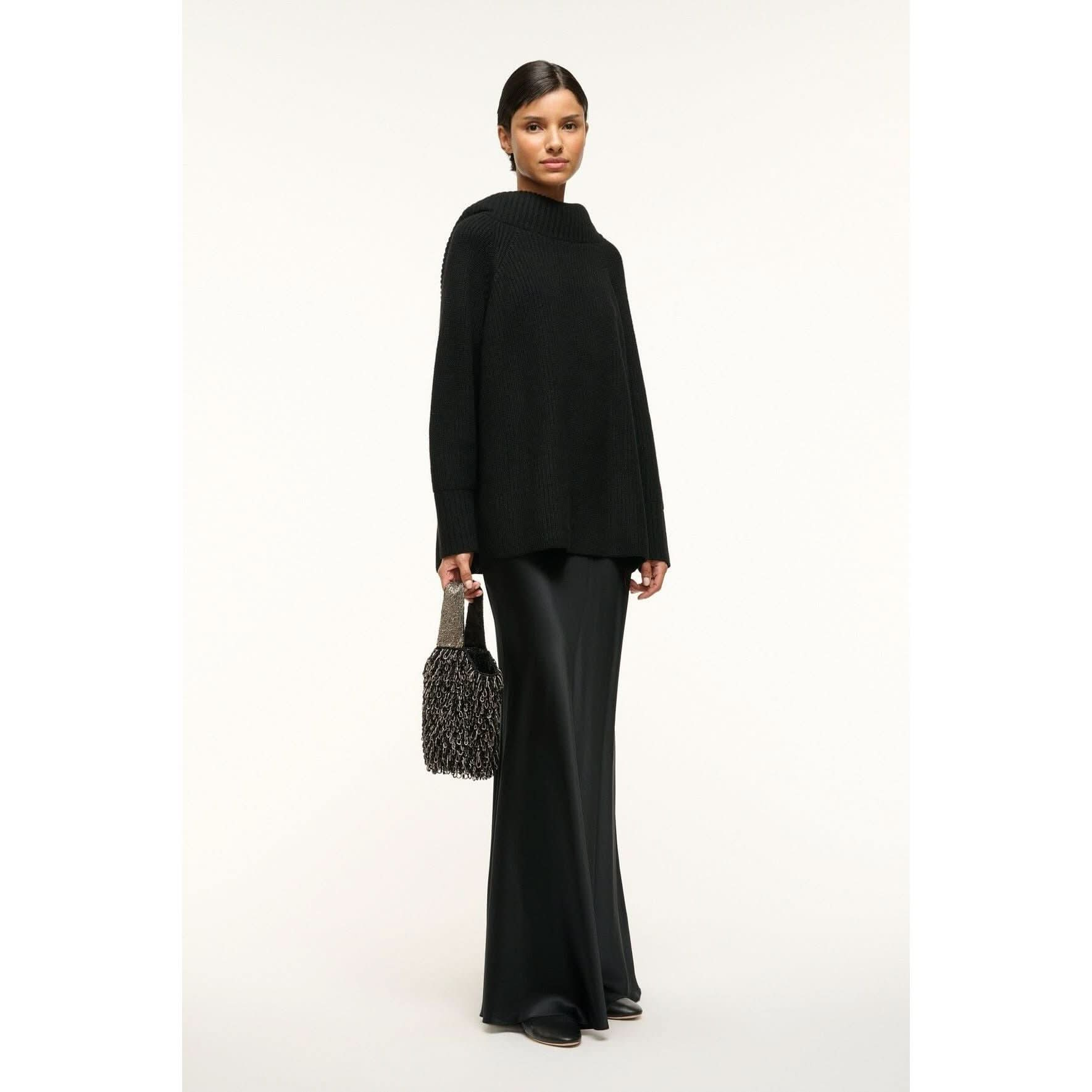 A person stands against a neutral background wearing a long black sweater and flowing black pants. They hold the Staud Haruko Beaded Bag in black, completing a stylish and minimalist look.