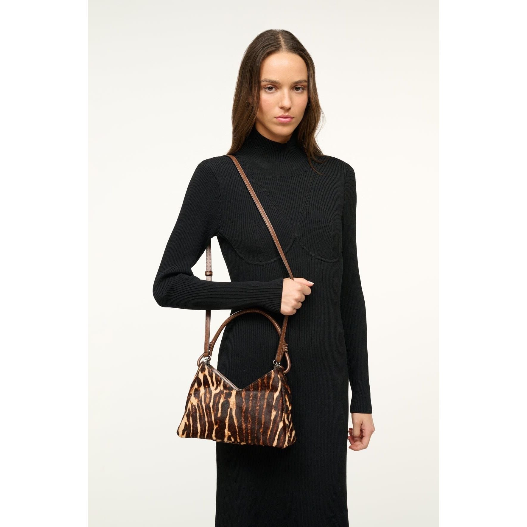 A person with long hair wears a black turtleneck dress and holds the Staud Valerie Shoulder Bag in Lady Leopard Cacao. This polished leather bag from Staud features a brown and black animal print design, adorned with two-tone hardware. It has two dark handles and is styled as a crossbody. The background is plain and light-colored.