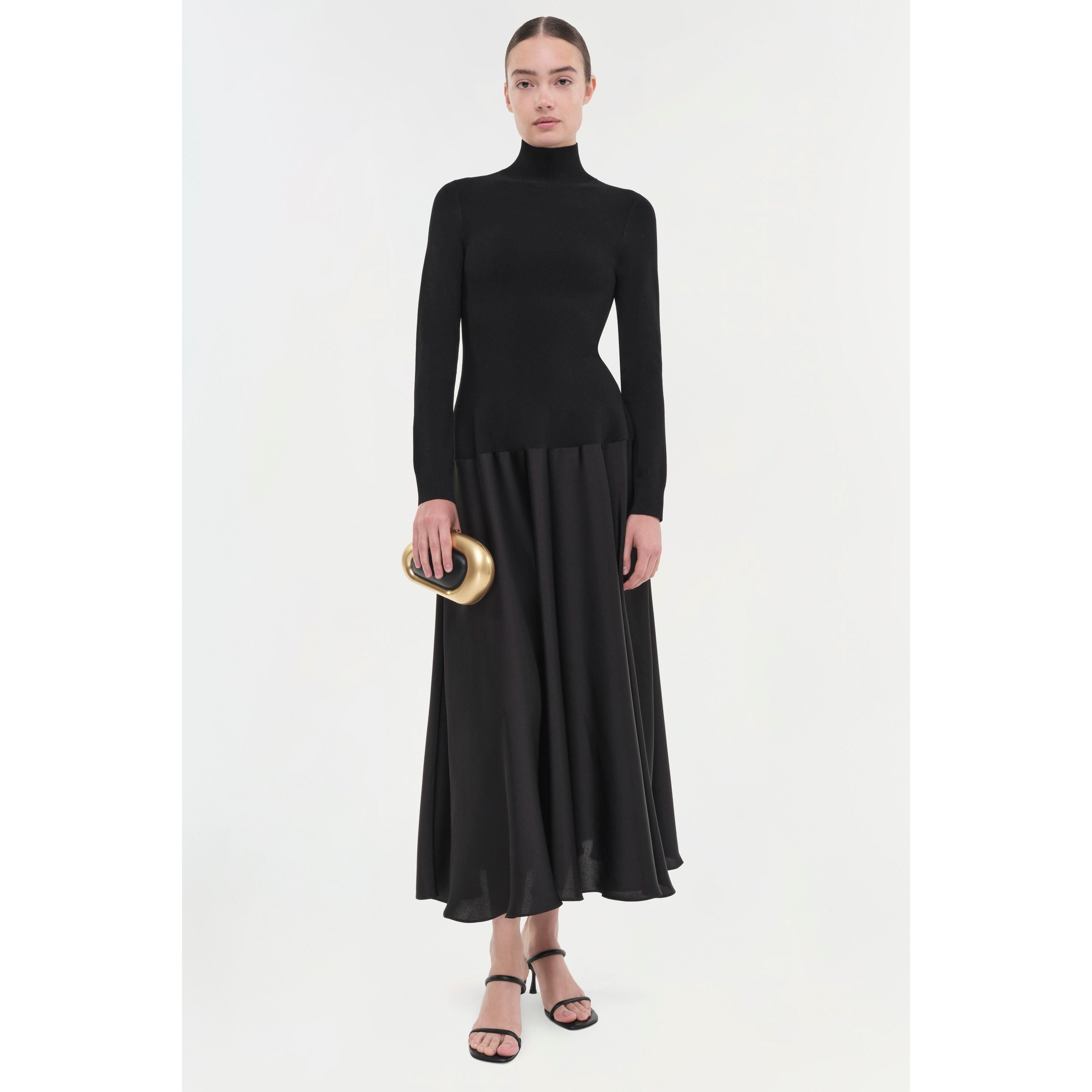 Dressed in a sleek Simkhai Frances Dress, a person stands gracefully holding a small, gold, heart-shaped object. The dress features a high neckline and pintucked waist, while their hair is neatly pulled back and they are wearing black sandals. The simple white background enhances the elegance of this ensemble by Simkhai.