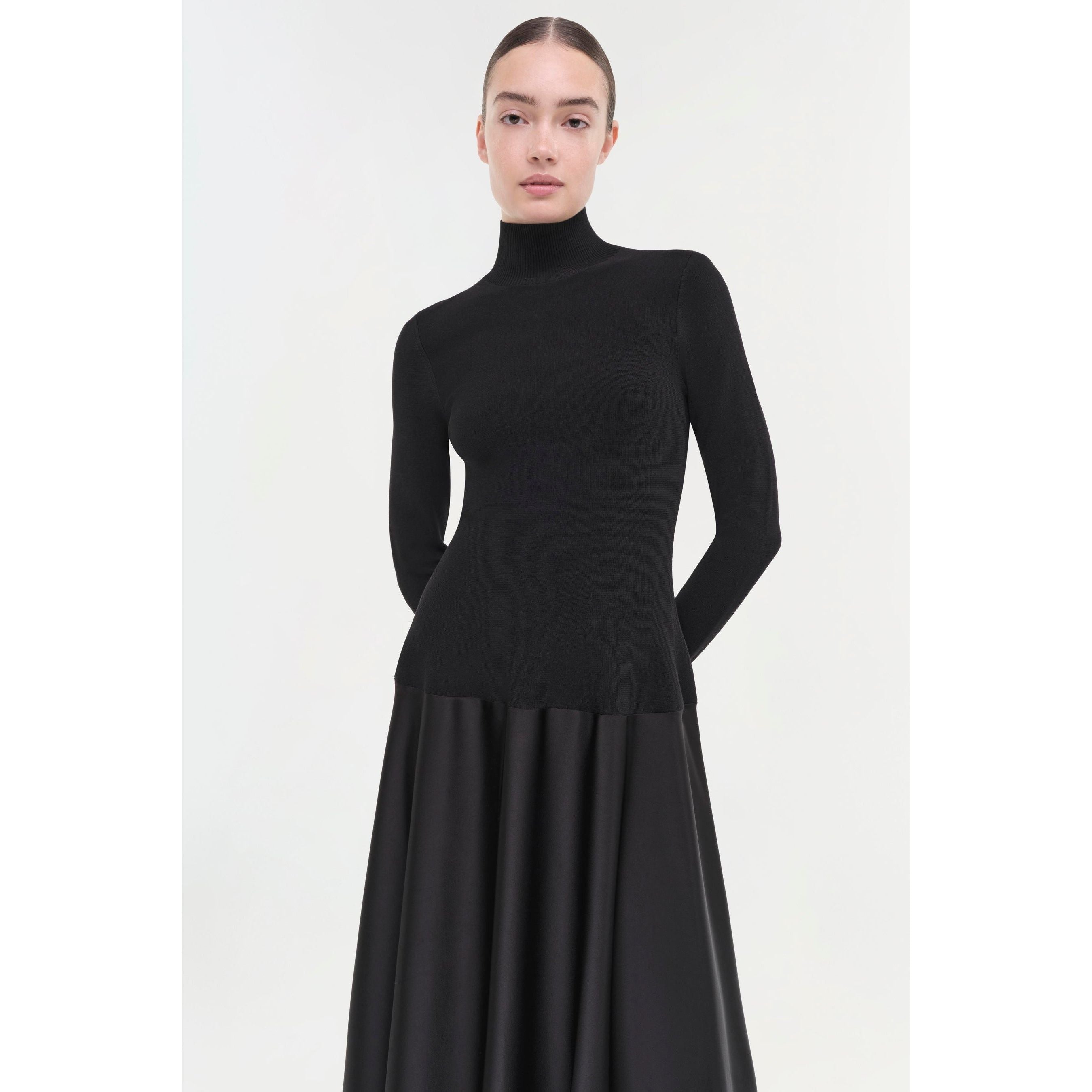 A woman wearing a black turtleneck top and the Simkhai Frances Dress, known for its pintucked waist and midi length, stands with her hands behind her back, looking forward against a plain white background.