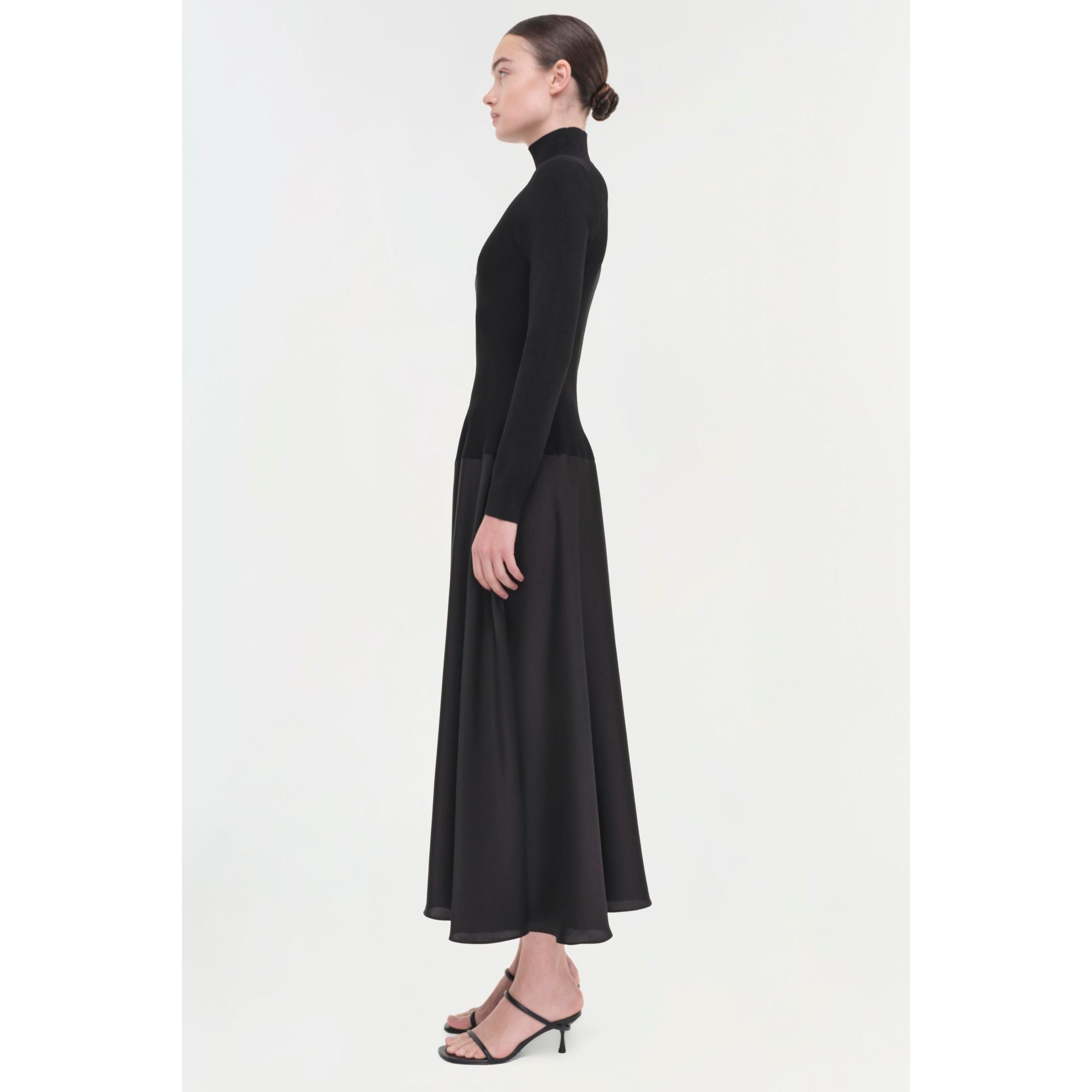 A person in a black Simkhai Frances Dress featuring a pintucked waist stands in profile against a plain background. The flowing midi-length skirt complements their black strappy heels, and their hair is pulled back neatly.