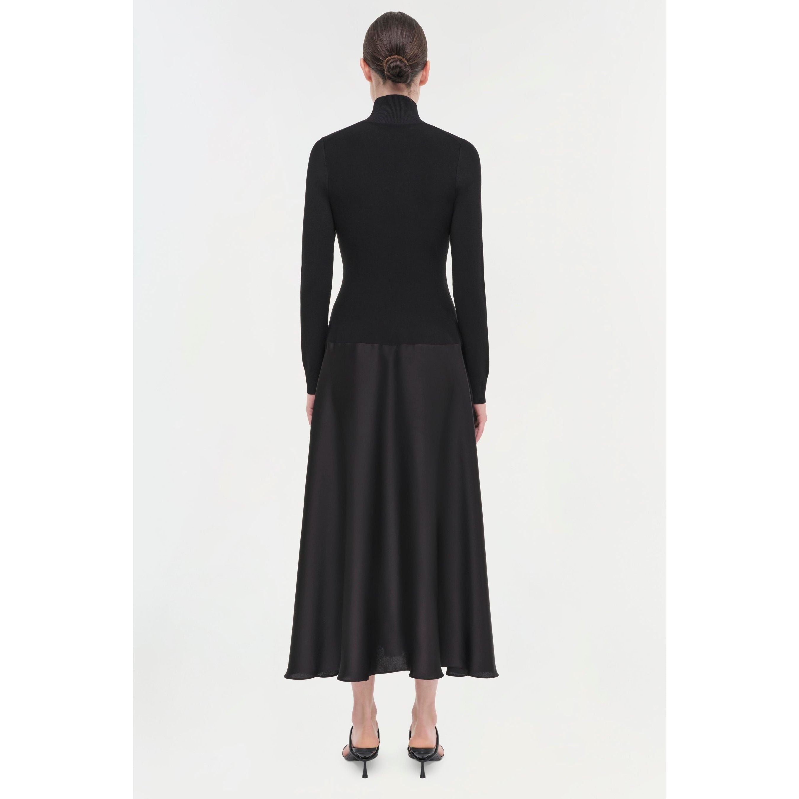 A woman with her hair in a bun faces away from the camera, displaying the elegant Simkhai Frances Dress. The ensemble's long-sleeved black design highlights a pintucked waist and is paired beautifully with black high-heeled sandals against a pristine white backdrop.