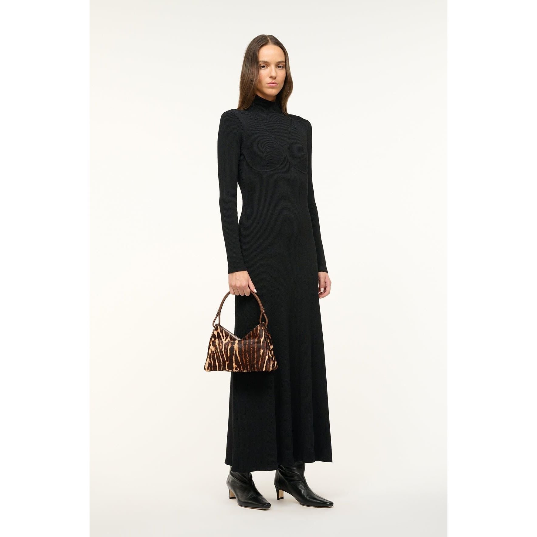 A person wearing a long black dress and black boots stands against a plain background, holding the chic Staud Valerie Shoulder Bag Lady Leopard Cacao.