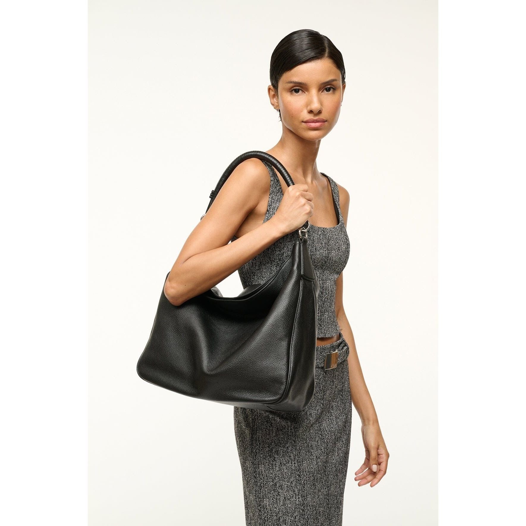 A person in a sleeveless gray dress carries a mid-size everyday bag, the Staud Perry Bag in black pebbled leather, elegantly resting on their shoulder against a plain, light-colored background.