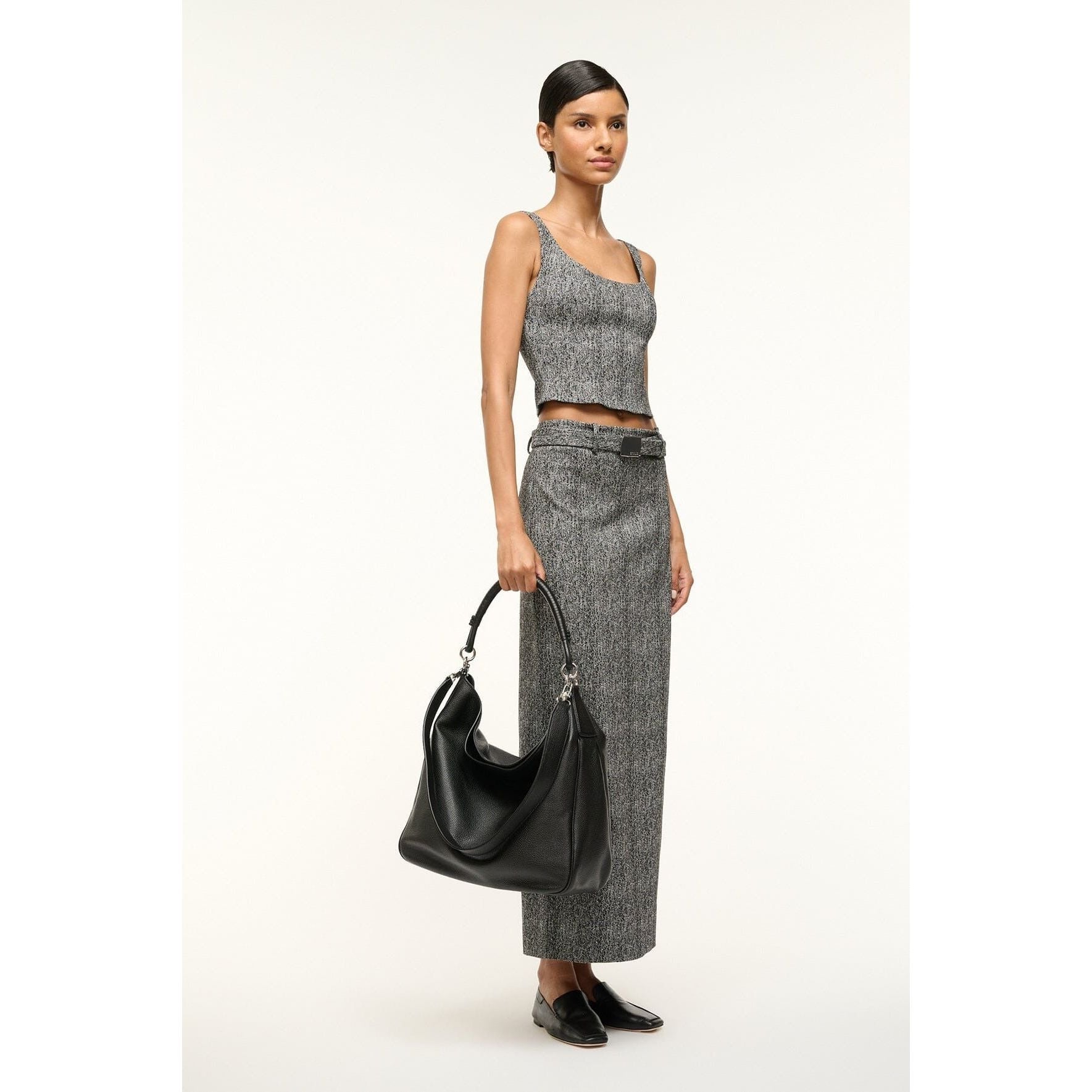 A model stands against a white background, wearing a gray sleeveless crop top paired with a matching long skirt. She carries the Staud Perry Bag Black, an everyday hobo shoulder bag crafted from pebbled leather. Her look is completed with black low-heeled shoes, and her hair is neatly styled as she exudes confidence.