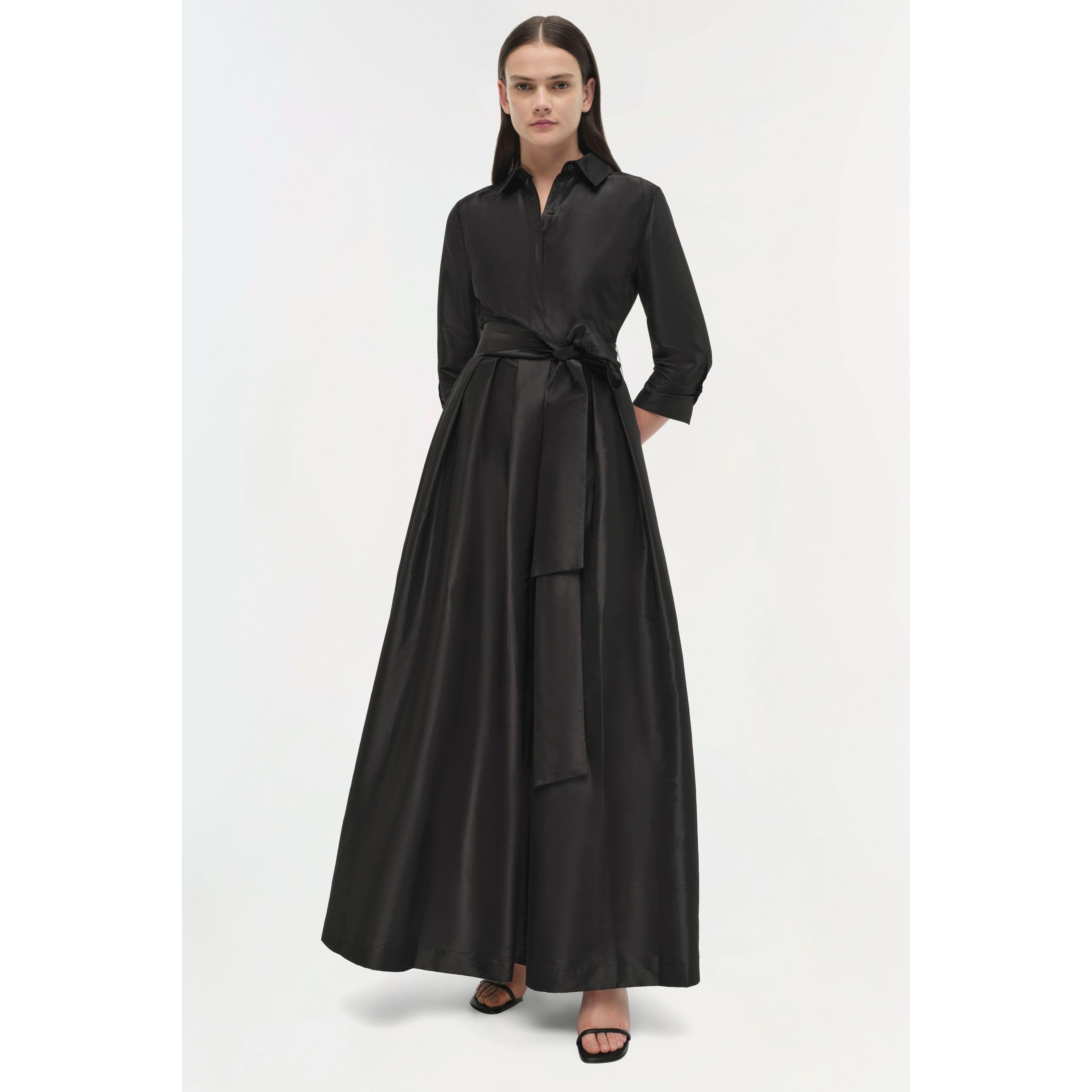 A person wearing a Simkhai Julip Belted Gown stands against a plain white background. The elegant black dress from Simkhai features a tied waist, long sleeves, and a structured shirt collar, gracefully flowing into a full-length skirt. Black open-toed sandals complete this sophisticated look.