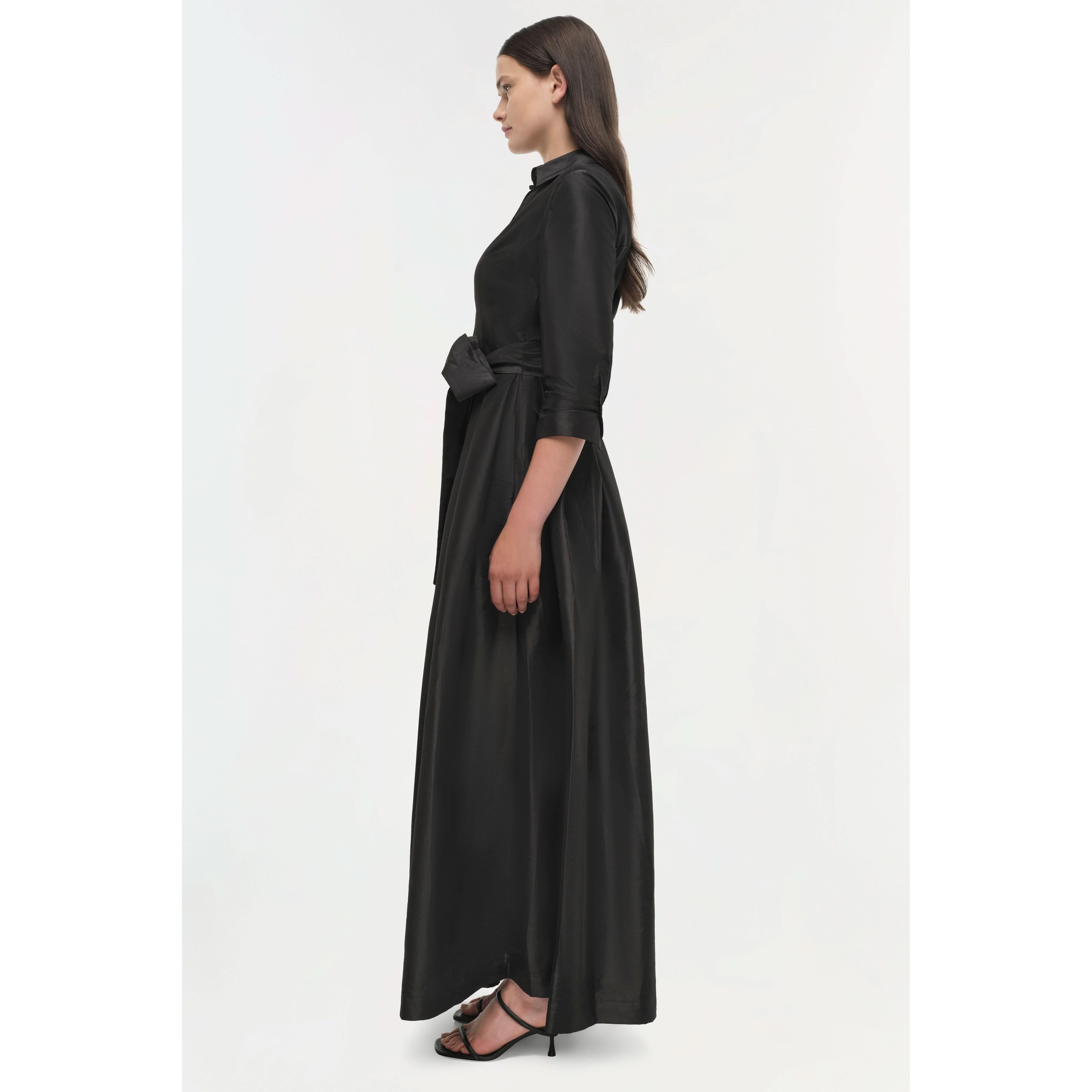 A woman in a Simkhai Julip Belted Gown, distinguished by its flowing full-length skirt and structured shirt collar, stands in profile. Her long brown hair cascades down as she wears black strappy heels, poised against a plain white background.