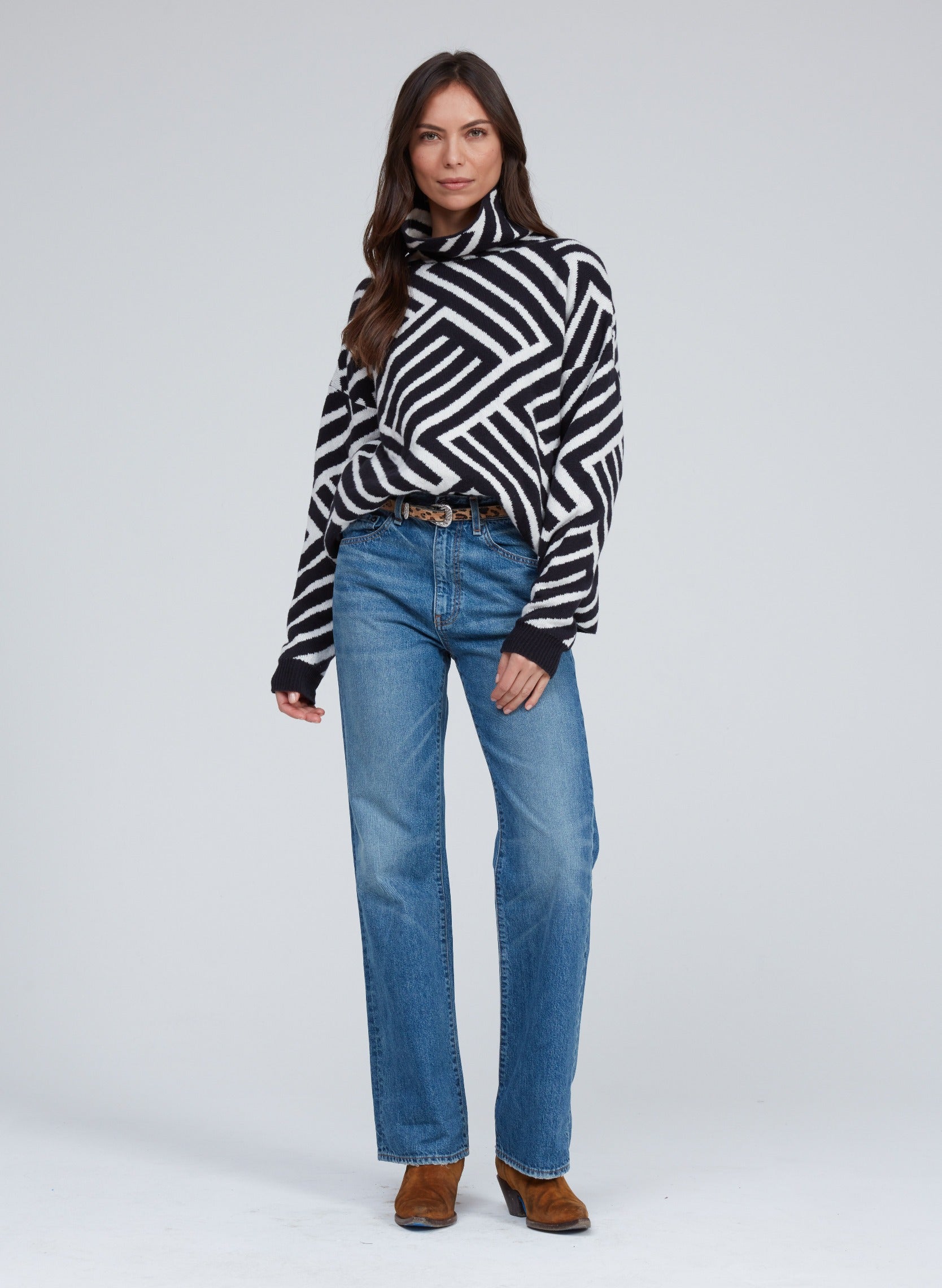 A woman stands against a plain background, wearing a black and white geometric-patterned turtleneck sweater, ASKK NY Straight Jean in Bush Baby wash by ASKK NY, and brown boots. She has long, wavy hair and is posed with her left hand in her pocket and her right hand relaxed at her side.