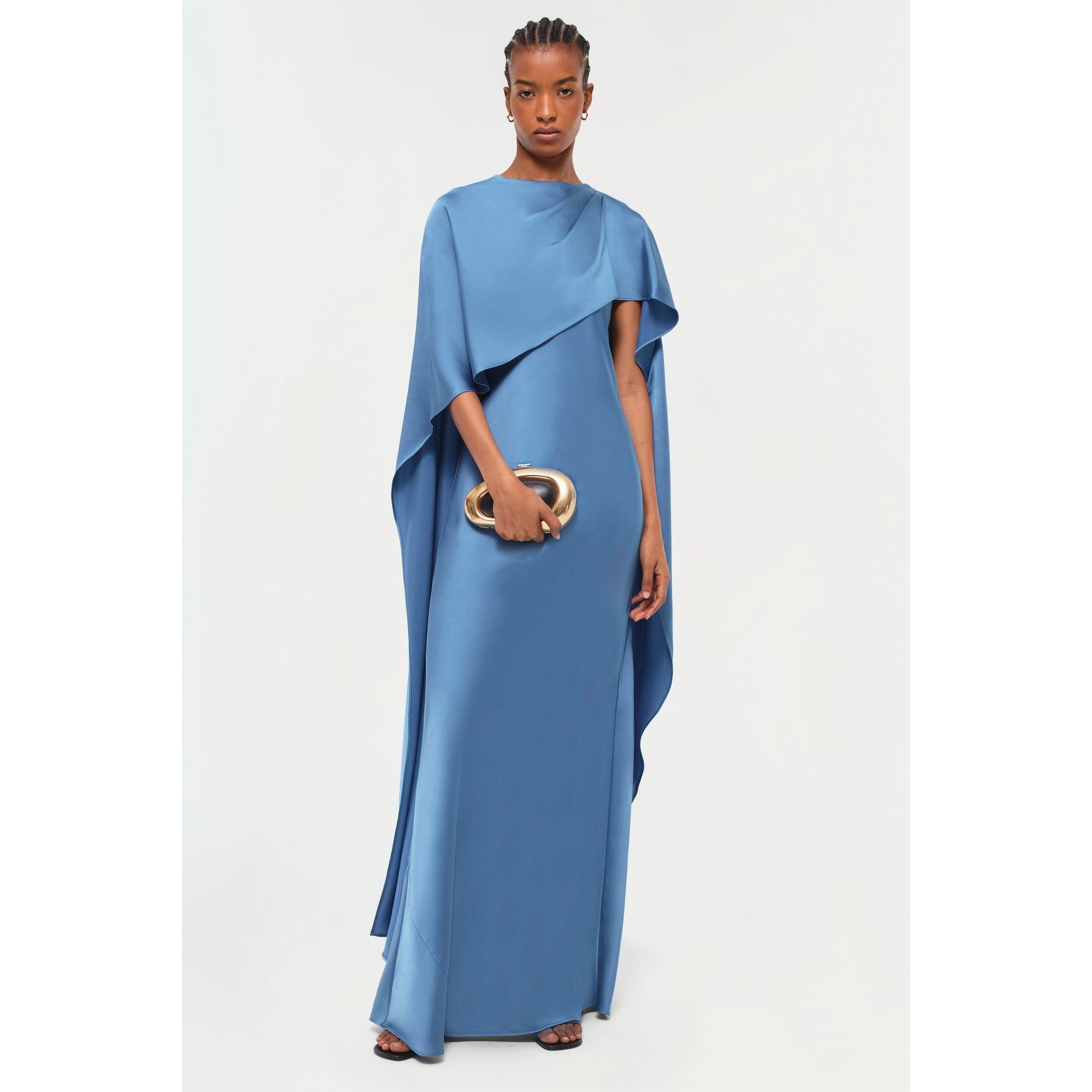 Dressed in the Simkhai Izel Cape Gown, a flowing blue ensemble with draped sleeves and a dramatic cape-like side, complemented by a bold front slit, an individual holds an elegant gold clutch. With neatly styled hair, they exude poise and confidence against a simple backdrop, as if showcasing an item from the Pre-Fall '24 Lookbook.