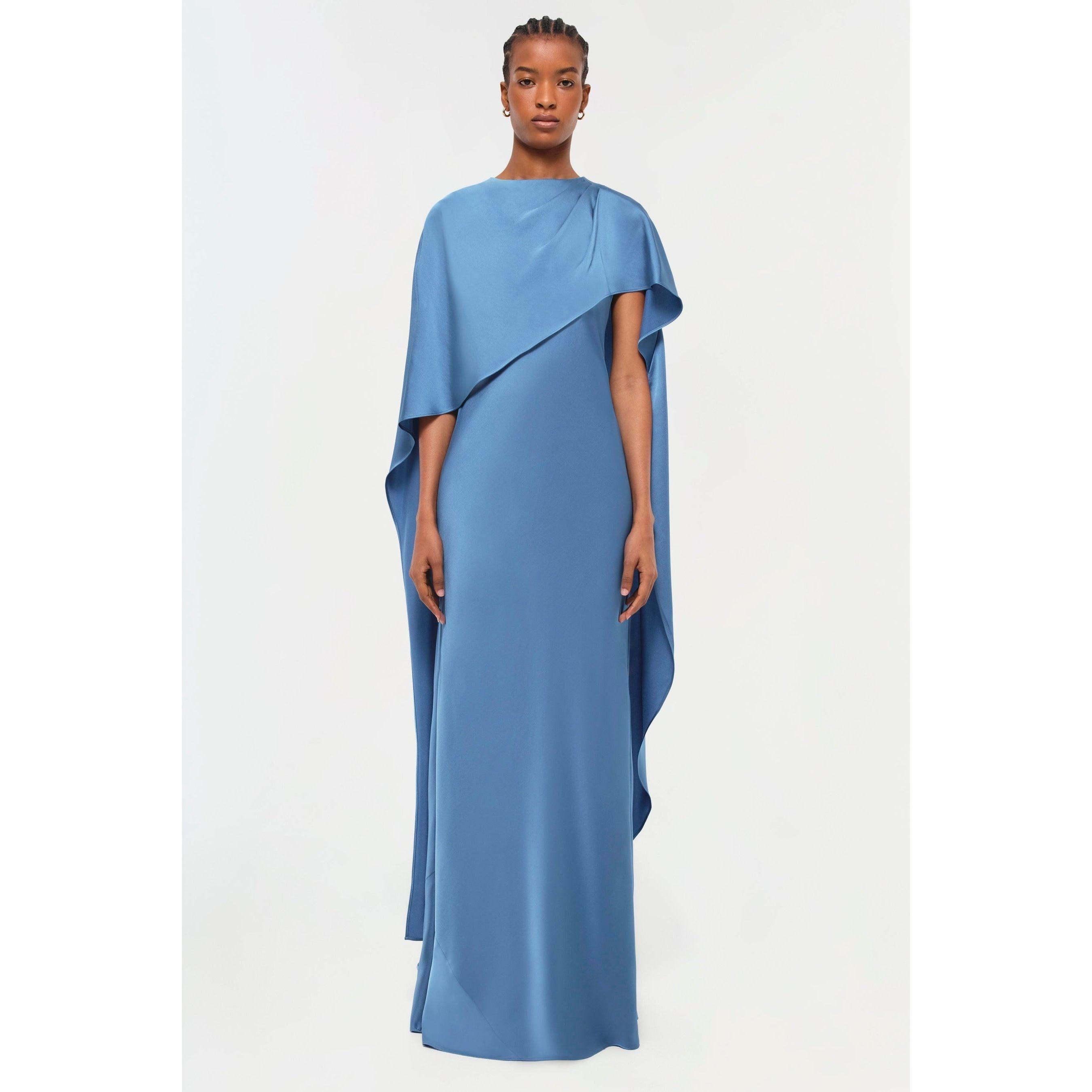 A person stands wearing the Simkhai Izel Cape Gown, a flowing asymmetrical blue dress with draped sleeves and a front slit, featured prominently in the Pre-Fall '24 Lookbook. The floor-length gown elegantly contrasts against the plain white background.