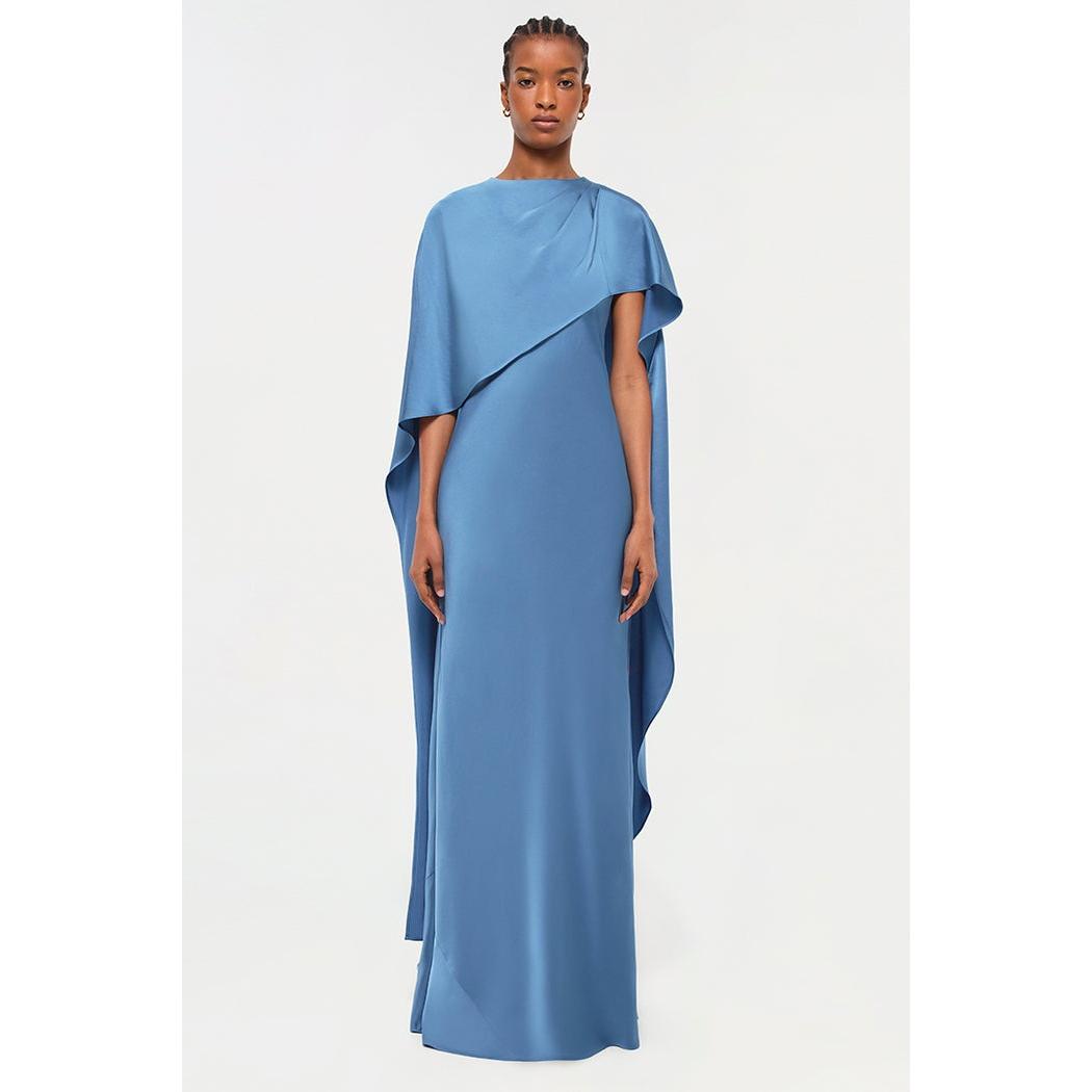 A person stands against a plain light background, wearing the Simkhai Izel Cape Gown from the Pre-Fall '24 Lookbook. This flowing blue gown features draped sleeves, a cape-like design over one shoulder, and a floor-length front slit. The person has short, styled hair and a calm expression.