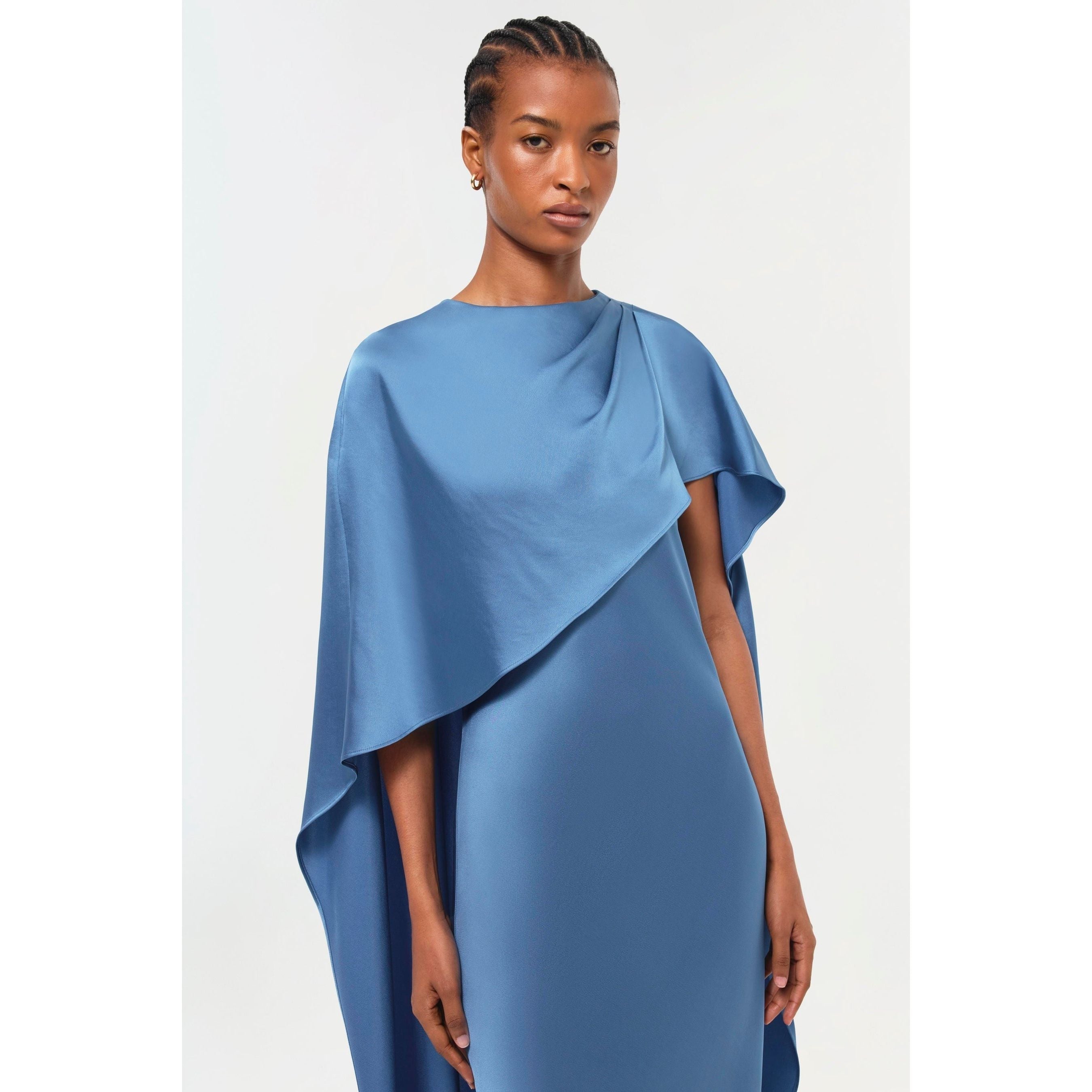 A person stands facing the camera, wearing the Simkhai Izel Cape Gown from the Pre-Fall '24 Lookbook. This elegant gown features draped sleeves and a cape-like design over one shoulder. The background is plain and light-colored.