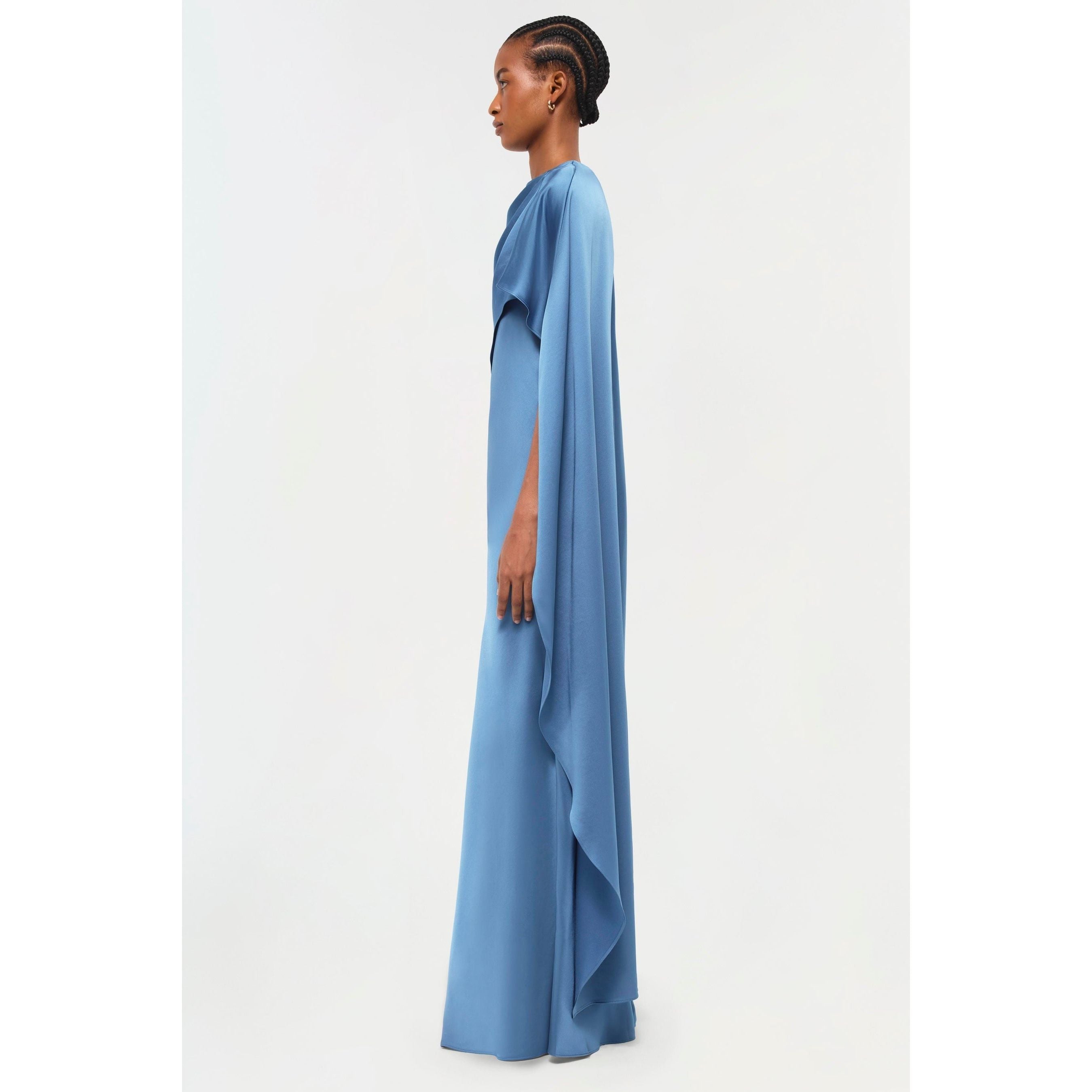 Against a simple backdrop, a person in profile elegantly displays the Simkhai Izel Cape Gown from the Pre-Fall '24 Lookbook. This graceful long blue gown features cape-like draped sleeves and a subtle front slit, complemented by their intricately braided hairstyle.