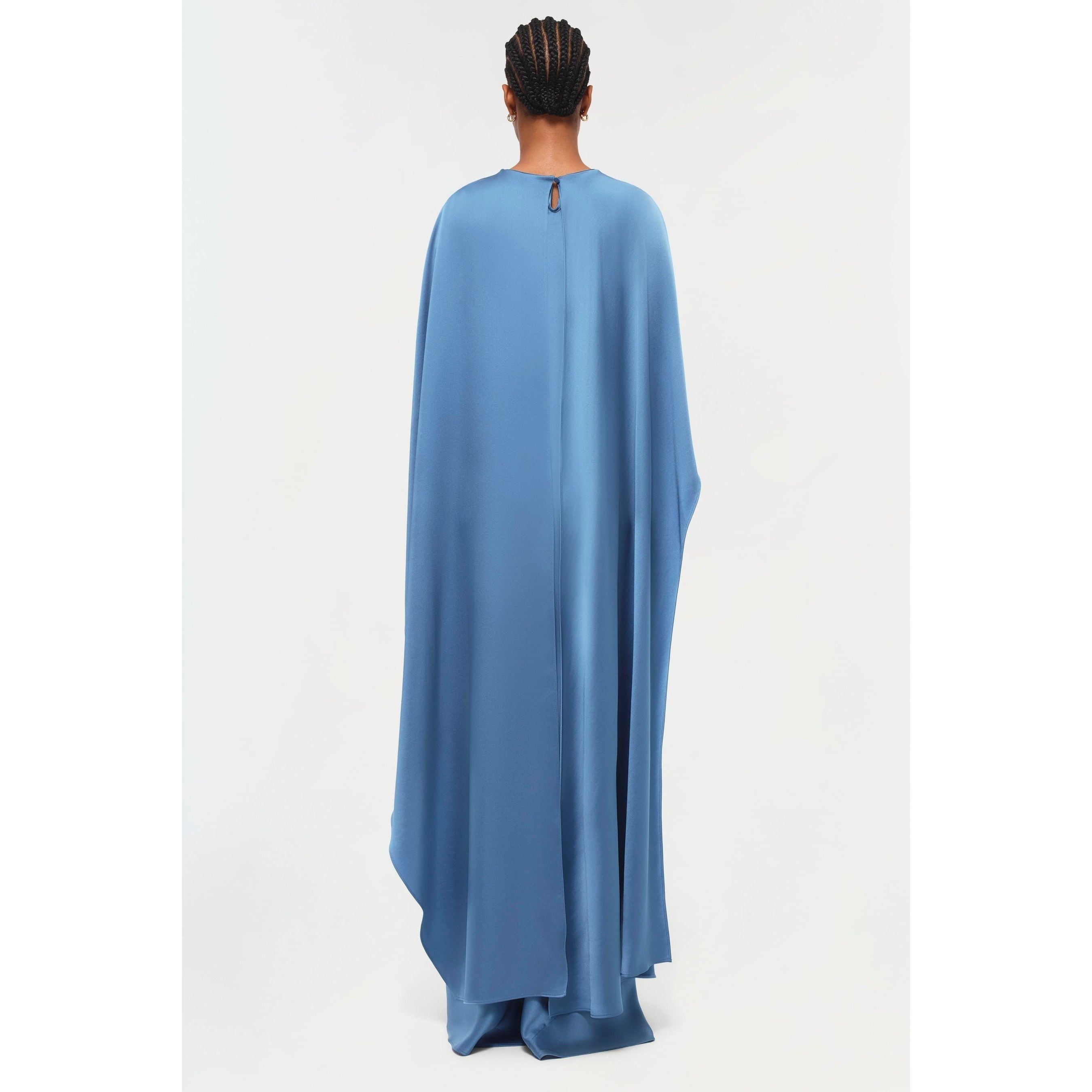 A person faces away from the camera, adorned in the Simkhai Izel Cape Gown, a stylish piece featured in the Pre-Fall '24 Lookbook. The gown's fabric elegantly cascades to the floor and is enhanced by beautifully draped sleeves. Their hair is styled in cornrows against a simple white backdrop.