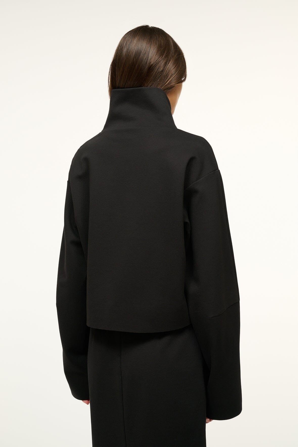 A person with long, dark hair is facing away from the camera, wearing the Staud Binoche Top in black, featuring a mock turtleneck and long sleeves over a matching black outfit. The plain, light-colored background highlights the top's details.