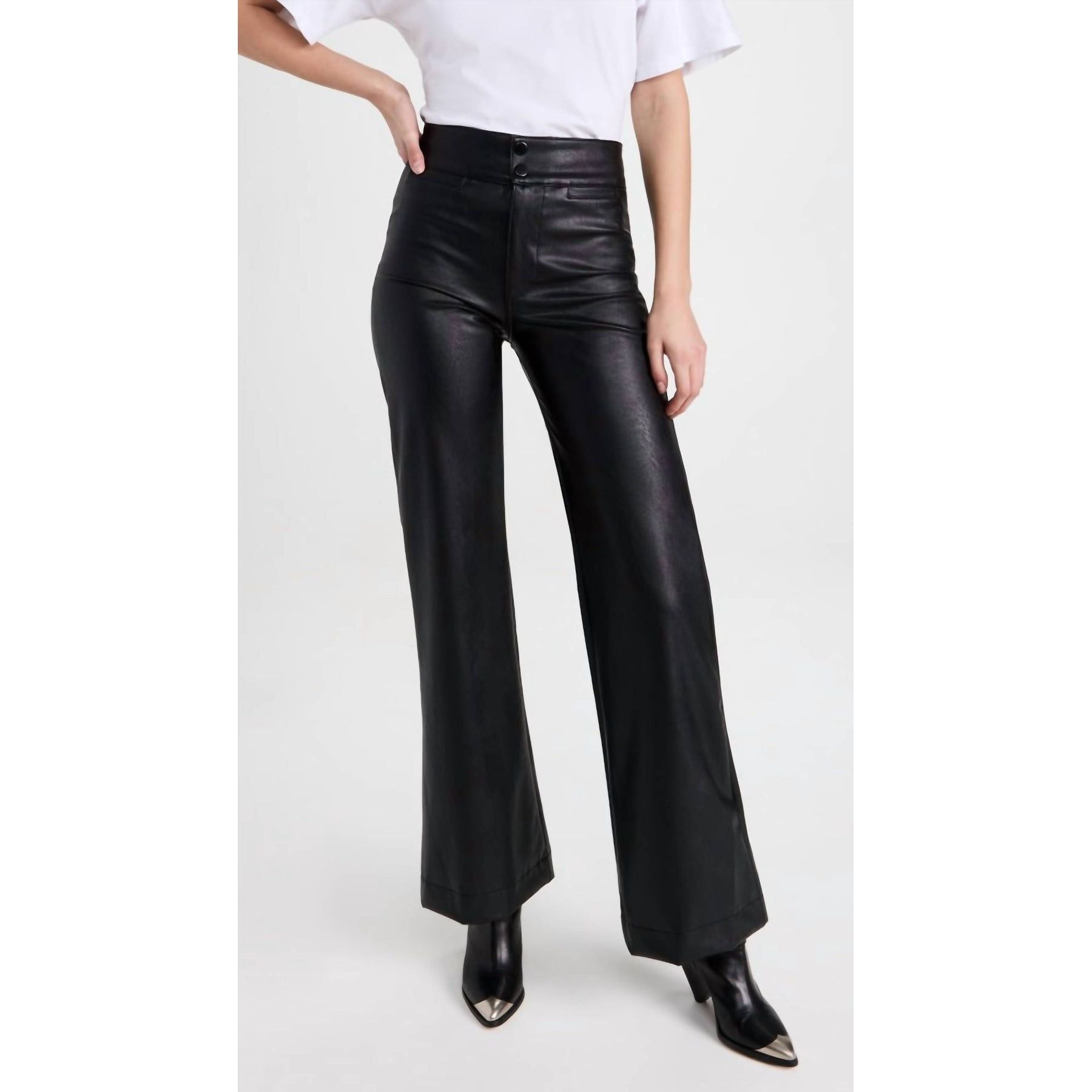 A person is wearing a white shirt paired with the ASKK NY Vegan Brighton Wide Leg Black, which offers the stylish appeal of classic wide-leg jeans, and black pointed-toe boots. Their left hand is resting on their hip against a light-colored background.