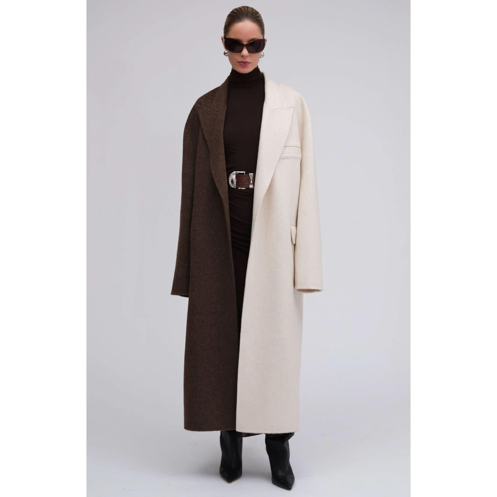 A woman stands confidently in a chic colorblocking ensemble, wearing the striking Ronny Kobo Wendalin Coat, which features one side brown and the other cream. She pairs it with a dark turtleneck, a stylish belt, black boots, and large sunglasses against a plain gray background.