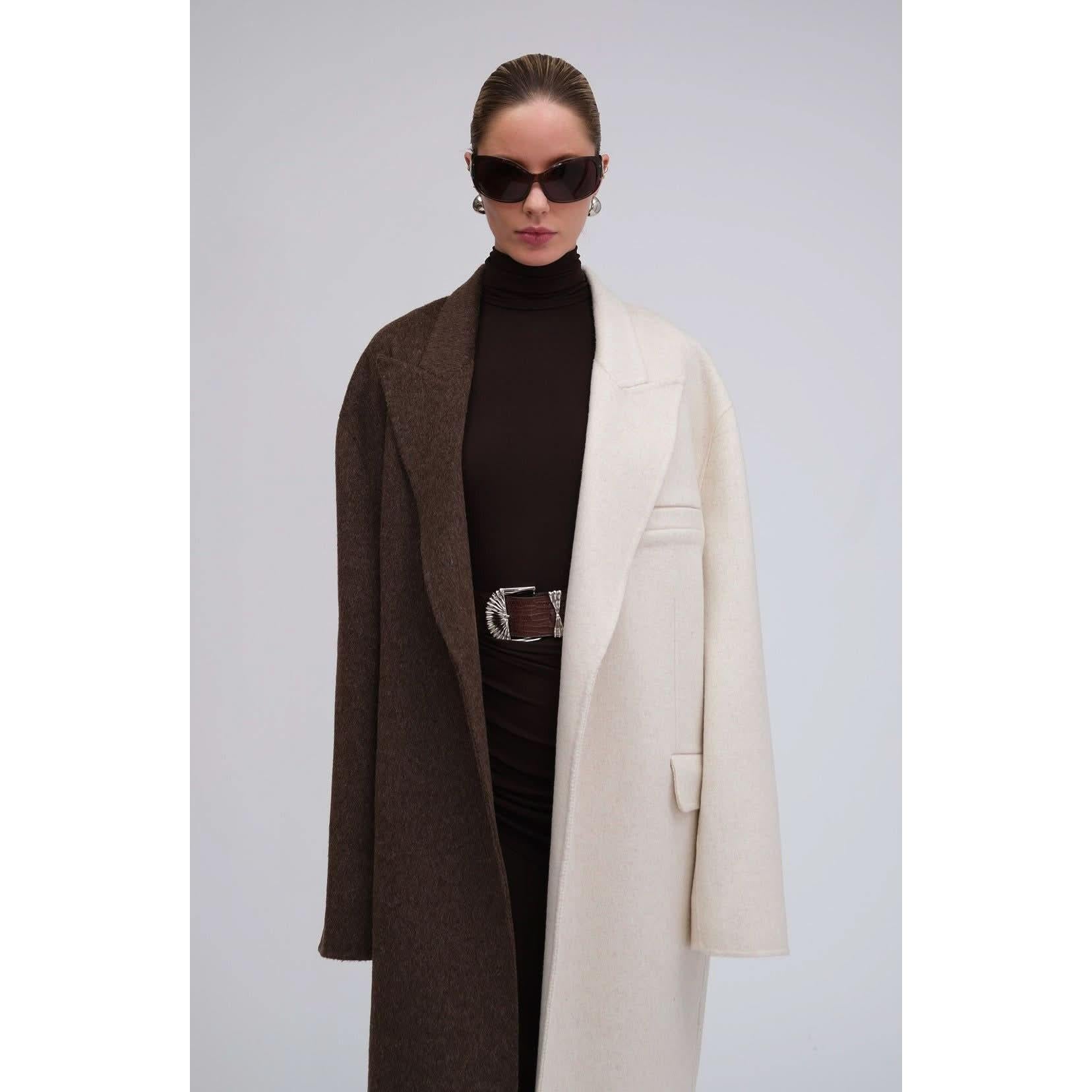 Positioned against a gray backdrop, an individual makes a statement in the Ronny Kobo Wendalin Coat, an oversized wool outfit with bold colorblocking featuring half-brown and half-cream tones. Complementing this look is a black turtleneck and dark sunglasses, while their pulled-back hair accentuates the silver-accented statement belt.