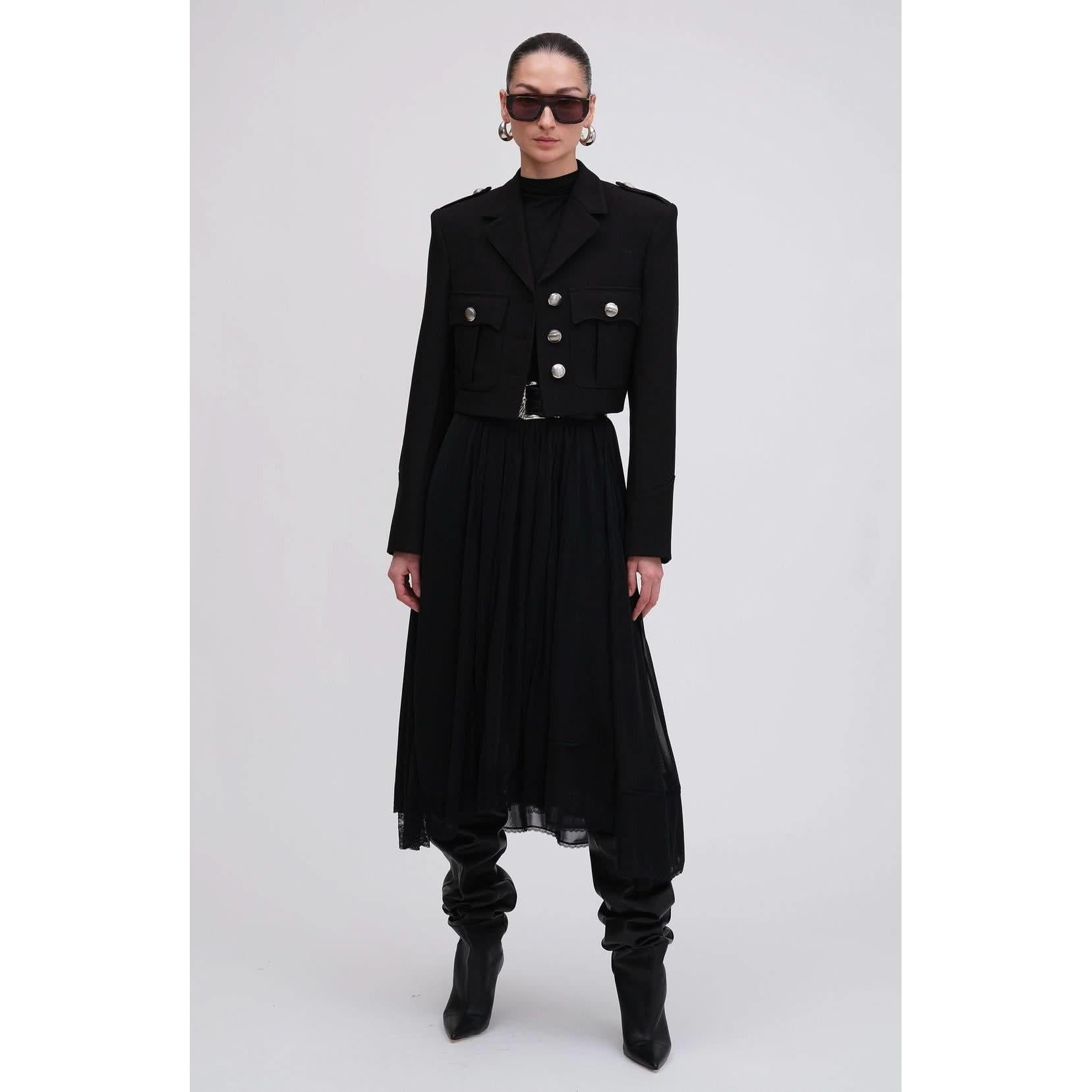 A person exudes Fall 2024 Style in an all-black outfit featuring the Ronny Kobo Genith Blazer with silver buttons, a long skirt, knee-high boots, and large sunglasses. The ensemble's sleekness is beautifully contrasted against the plain white background.