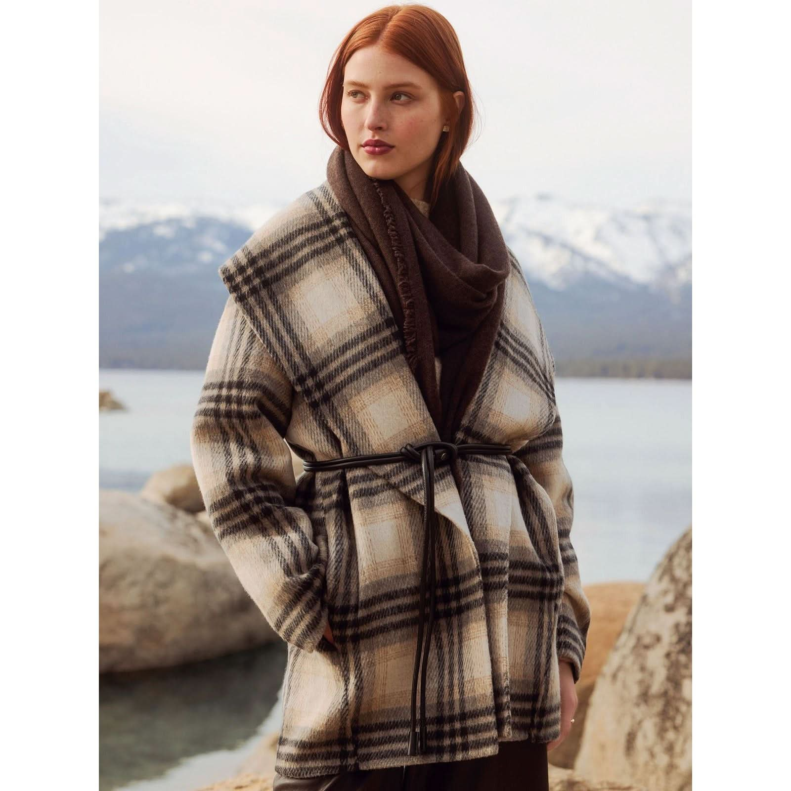 A woman with long, auburn hair is standing outdoors near a lake and mountains. She is wearing the Brochu Walker The Finley Plaid Coat in a relaxed fit, woven from cotton wool slub yarn. The background features rocks, water, and snow-capped mountains under a clear sky.