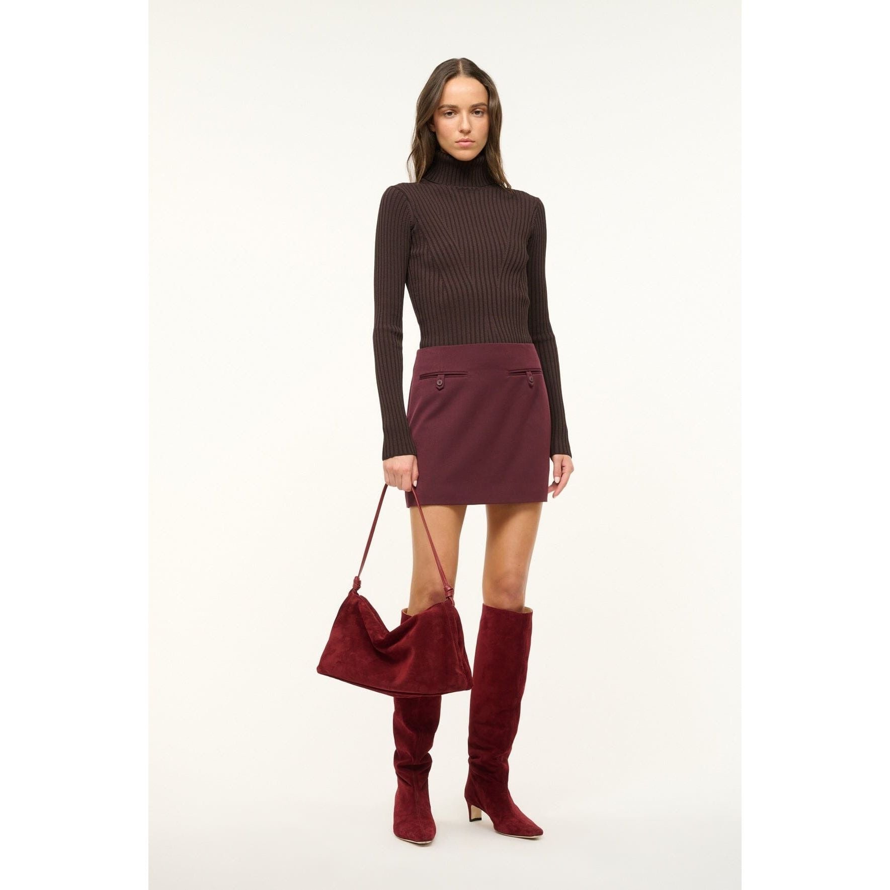 A person dressed in a dark ribbed turtleneck and a burgundy skirt is holding the fashionable Staud Wally Bag Pinot. They are also wearing knee-high burgundy boots, which perfectly complement their ensemble. The plain, light-colored background accentuates their chic appearance.