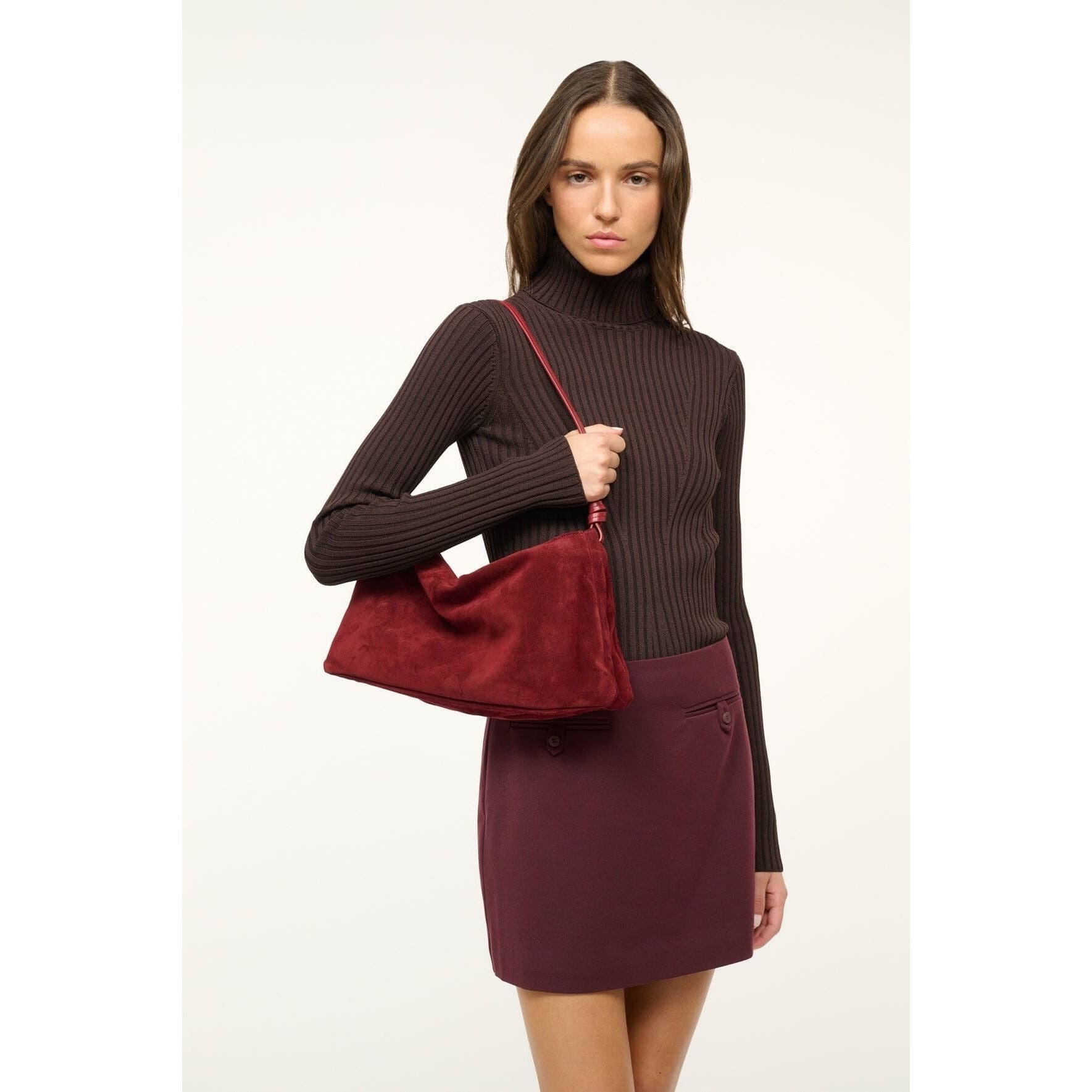 A person with long hair wears a dark ribbed turtleneck and a maroon skirt, holding a Staud Wally Bag Pinot over their shoulder. They stand against a plain white background.