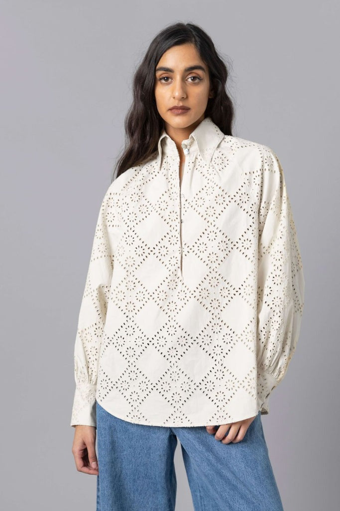 A woman with long dark hair wears the Hope for Flowers Pull Over Shirt Ecru by Hope for Flowers by Tracy Reece—a white, eyelet-patterned blouse with a collar and long sleeves made from GOTS certified organic cotton. She pairs it with light blue jeans in front of a plain gray background.