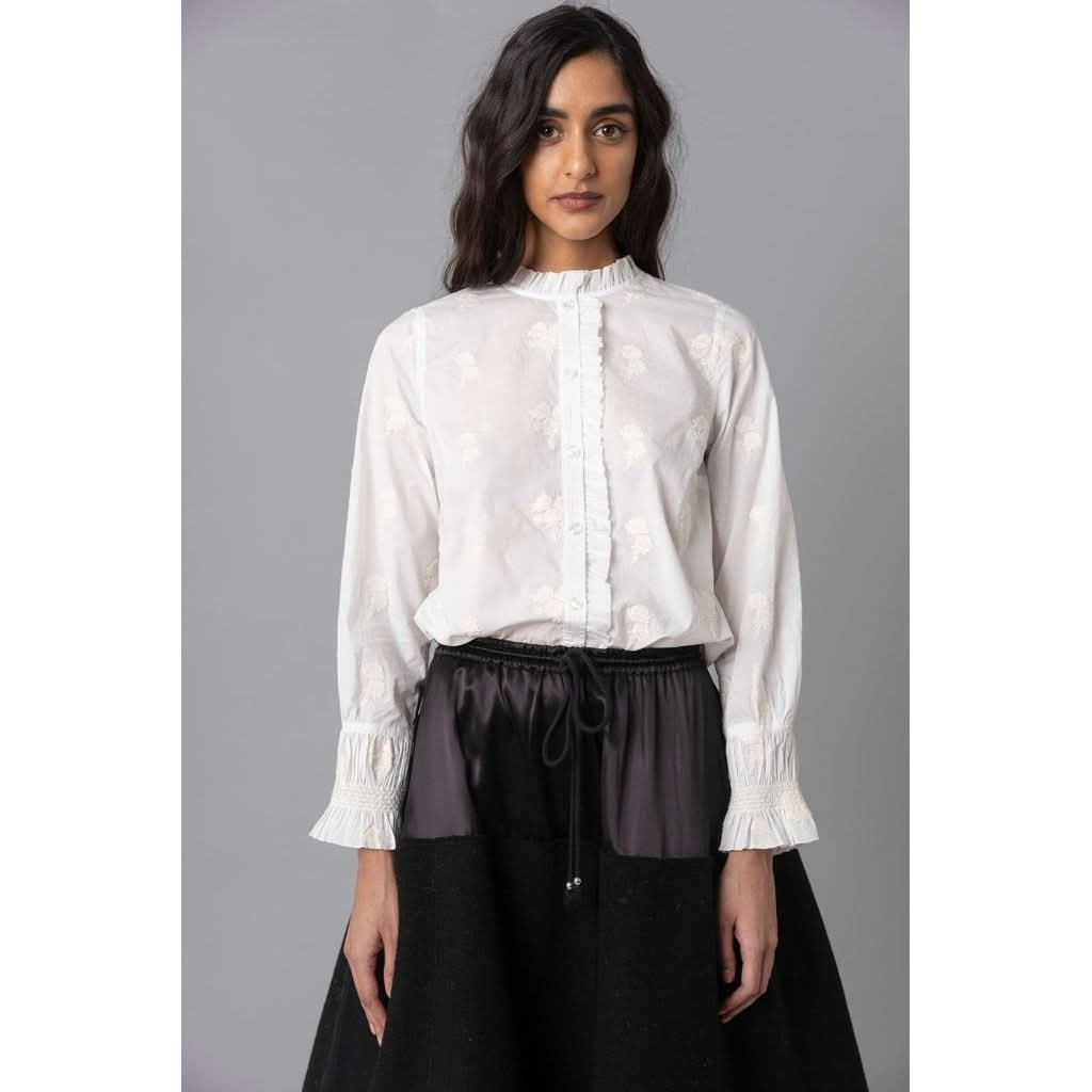 A woman with long dark hair is wearing the Hope for Flowers Petite Fleur Shirt by Hope for Flowers by Tracy Reece, which features intricate detailing and ruffled cuffs. She has paired it with a black skirt with a gathered waist. The ensemble reflects ethical manufacturing, set against a plain gray background.