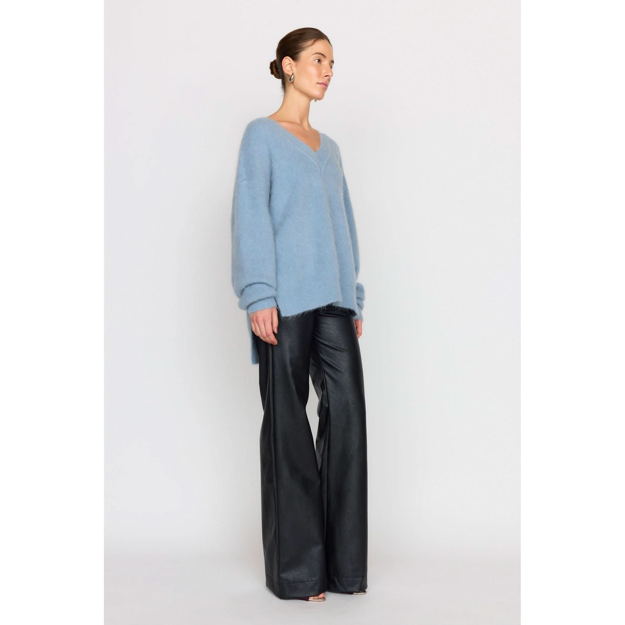 A woman styles a Christy Lynn Allie Sweater in Dusty Blue with black leather wide-legged pants against a plain background. Her hair is tied back, accentuating her hoop earrings.