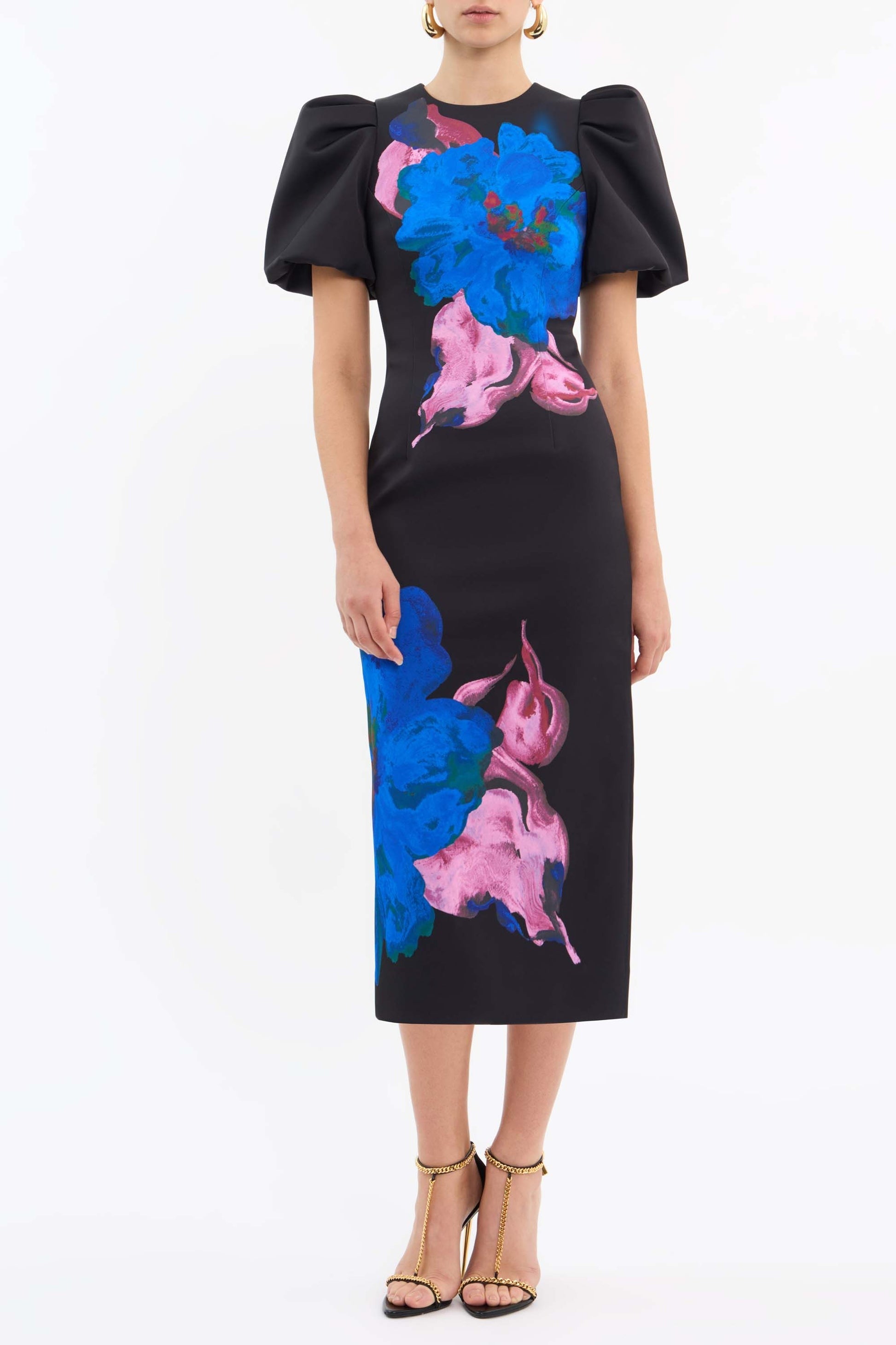 A woman models the Rebecca Vallance Acid Bloom Midi, a black midi dress by Rebecca Vallance with a fitted bodice, puff sleeves, and colorful floral prints in blue and pink. She pairs it with gold heels and stands against a plain white background.