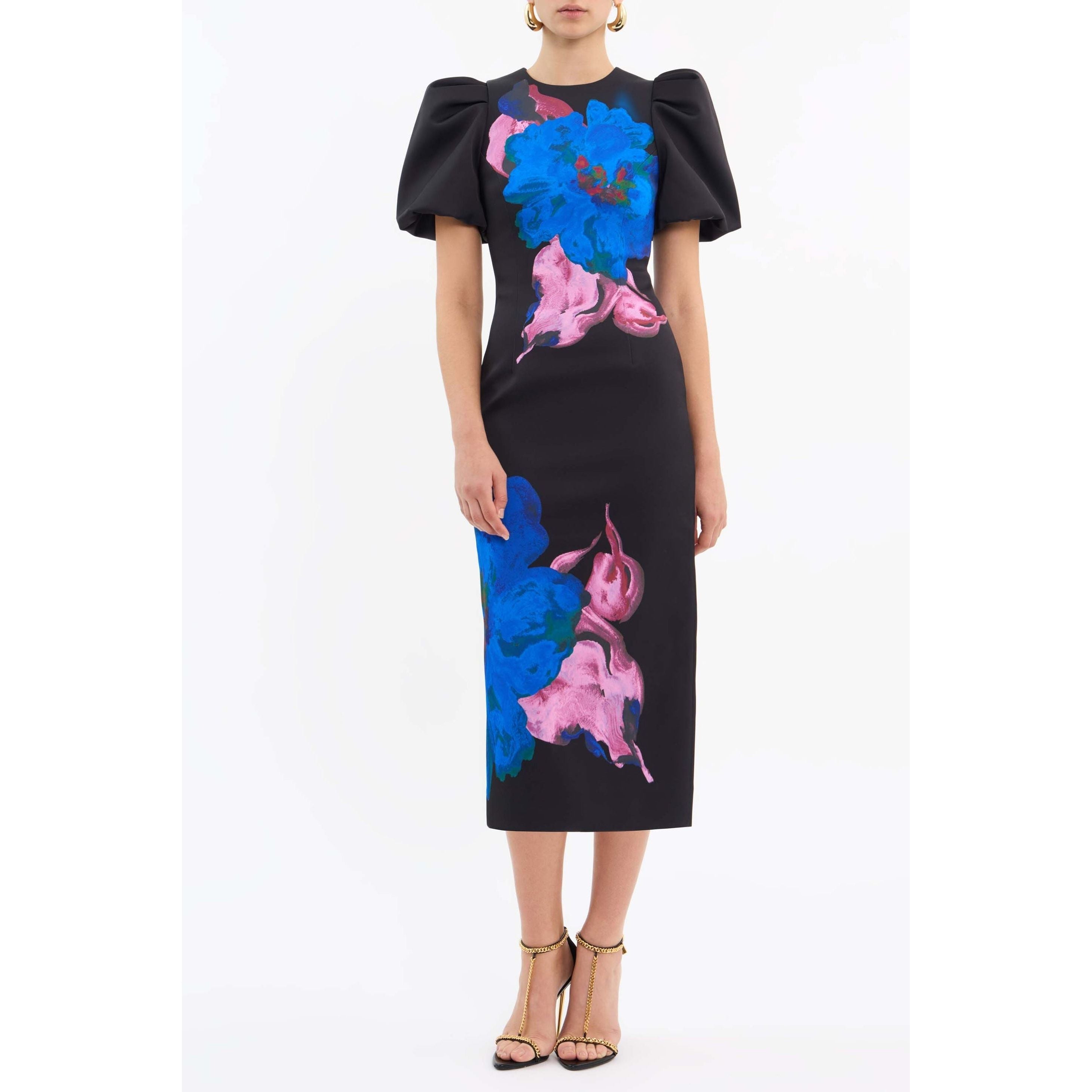 A woman models the Rebecca Vallance Acid Bloom Midi, a black midi dress by Rebecca Vallance with a fitted bodice, puff sleeves, and colorful floral prints in blue and pink. She pairs it with gold heels and stands against a plain white background.