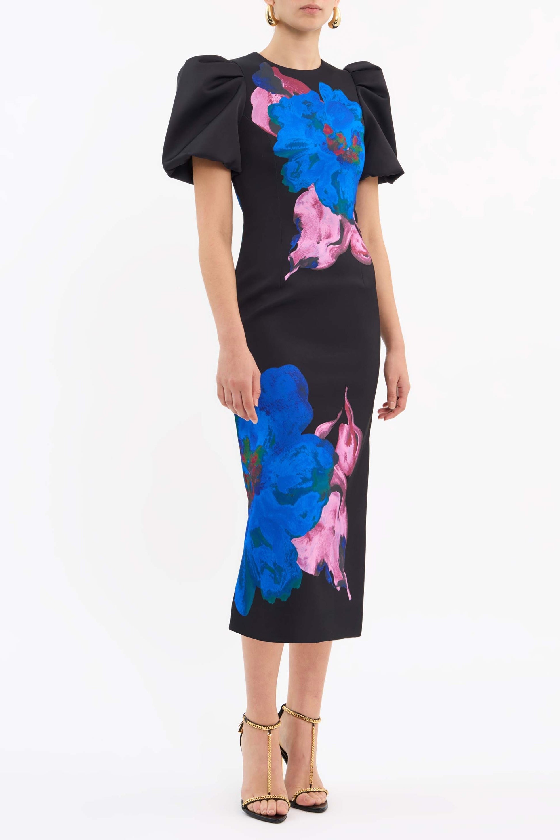 A person is wearing the Rebecca Vallance Acid Bloom Midi dress, which showcases a fitted bodice and short, puffed sleeves adorned with large blue and pink floral prints against a black background. Complementing the look, they are wearing gold strappy high-heeled sandals, all set against a plain white backdrop.