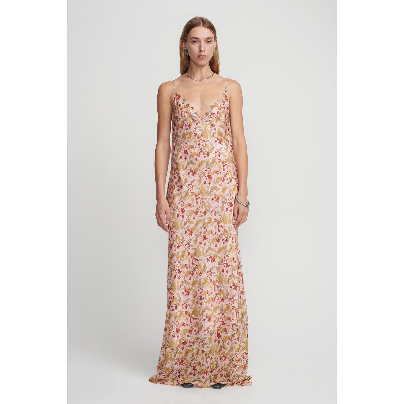 A person with long, straight hair is wearing the Hansen & Gretel Aggie Maxi Dress featuring a delicate floral pattern in shades of red, green, and gold on a light-colored background. The halter neck design by Hansen & Gretel adds an elegant touch as they stand facing the camera against a plain white backdrop.