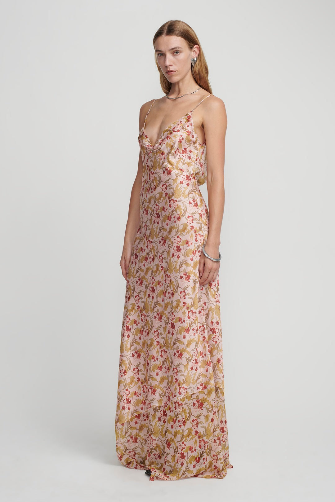 A woman with long, straight hair is wearing the Hansen & Gretel Aggie Maxi Dress, a floor-length floral gown with thin straps, as she stands against a plain background. She has a neutral expression and wears minimal jewelry, including earrings and a bracelet.