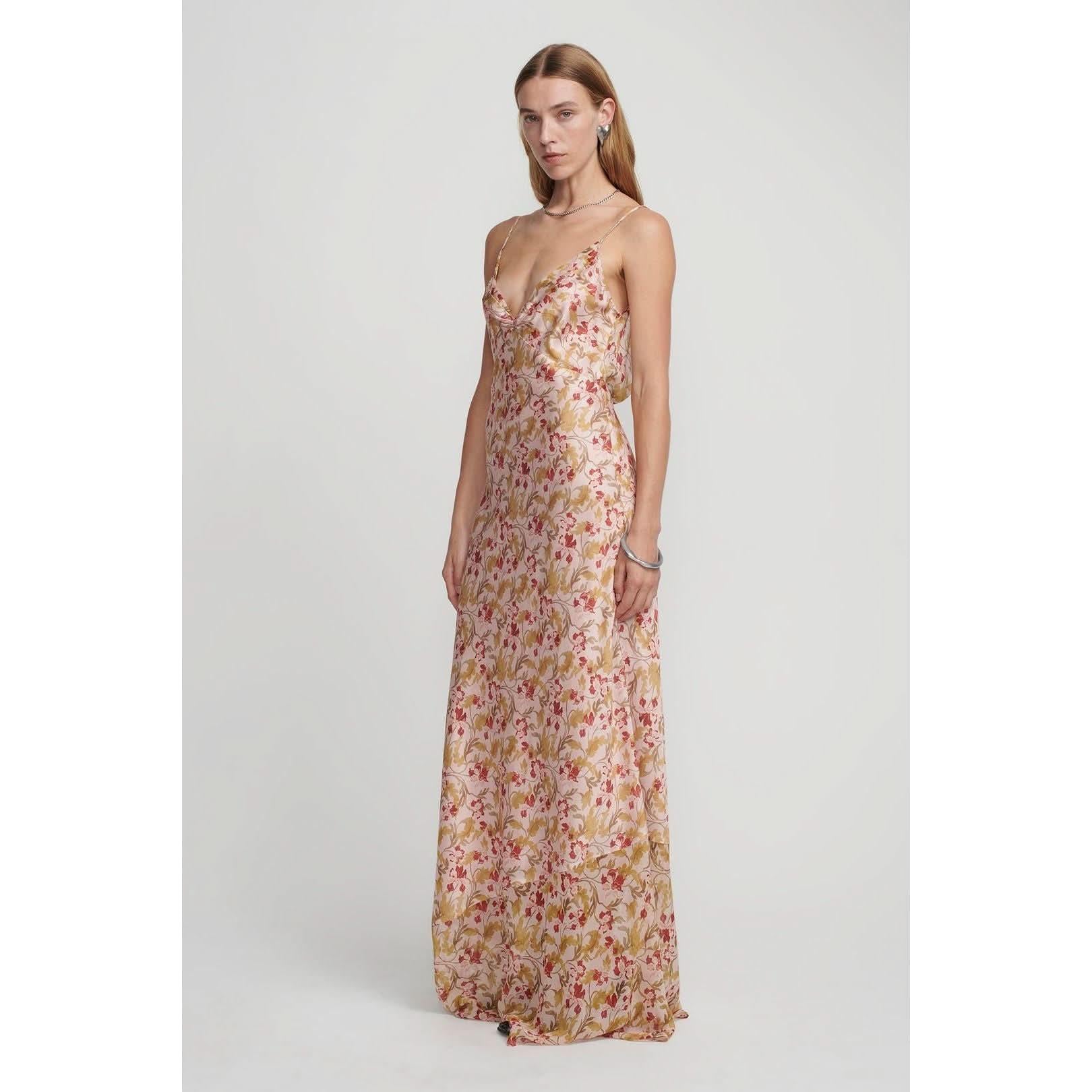 A woman with long, straight hair is wearing the Hansen & Gretel Aggie Maxi Dress, a floor-length floral gown with thin straps, as she stands against a plain background. She has a neutral expression and wears minimal jewelry, including earrings and a bracelet.