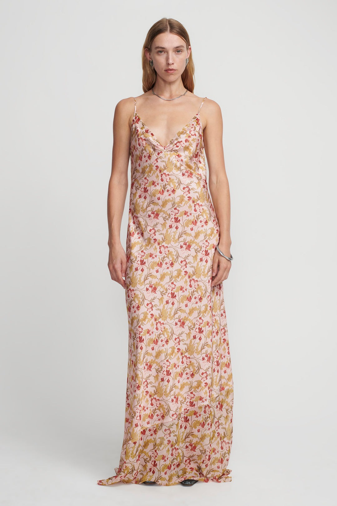 A person stands against a plain background, wearing the Hansen & Gretel Aggie Maxi Dress. The long, sleeveless gown showcases a beautiful floral print in shades of pink, red, and green. The dress features thin straps and a V-neckline. The person has long, straight hair and a neutral expression.