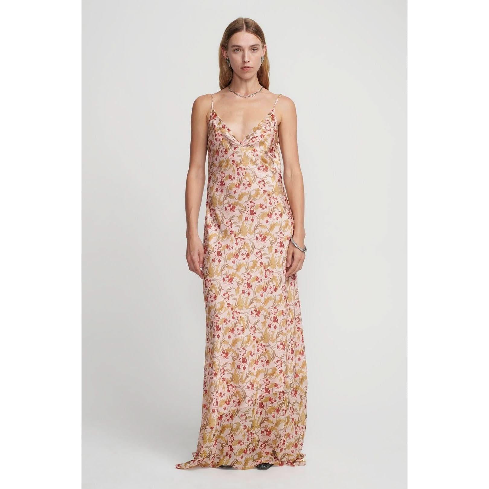 A person stands against a plain background, wearing the Hansen & Gretel Aggie Maxi Dress. The long, sleeveless gown showcases a beautiful floral print in shades of pink, red, and green. The dress features thin straps and a V-neckline. The person has long, straight hair and a neutral expression.