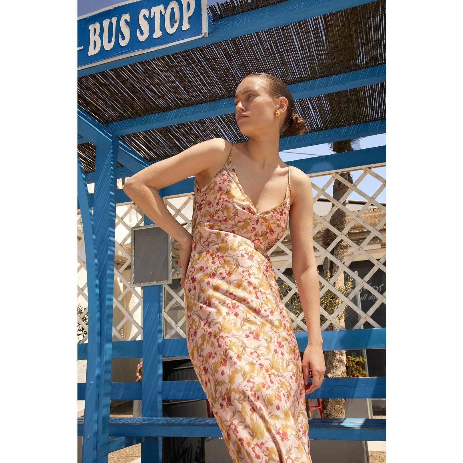 A person stands in front of a blue bus stop shelter on a sunny day, wearing the Hansen & Gretel Aggie Maxi Dress. They have their hand placed on their hip. The bus stop features a woven roof and a white lattice design in the background.