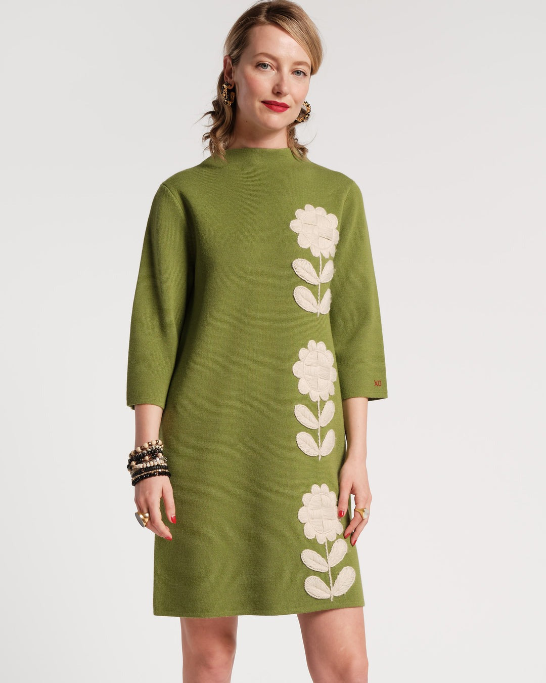 A person with shoulder-length hair stands with their back to the camera, wearing a green Frances Valentine Annie Dress Posy Embroidery Merino Wool Fern. The knee-length dress features three-quarter sleeves and is paired with black flats. Several bracelets adorn the person’s wrist, and they are positioned against a plain white background.